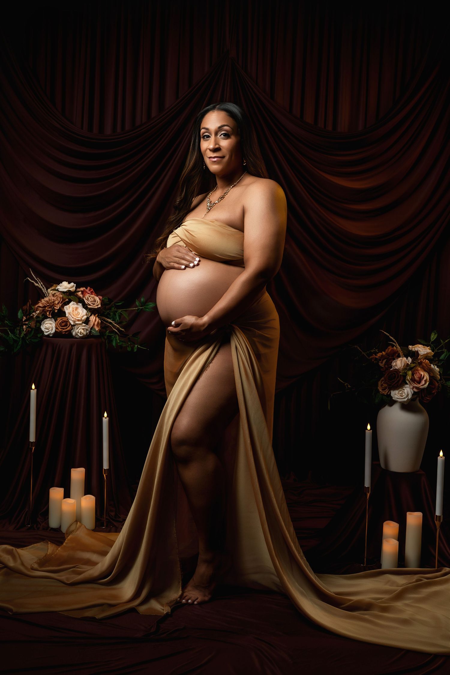 Moody maternity portrait with brown draping and candlelit setting featuring floral arrangements.