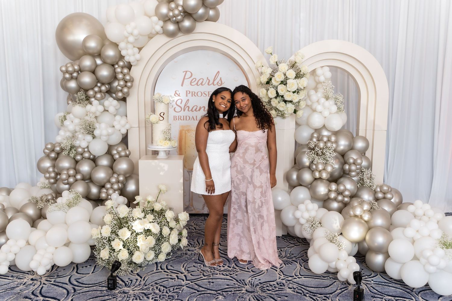 Elegant photo backdrop with silver and white balloon garland arrangements and floral decorations against a white wall.