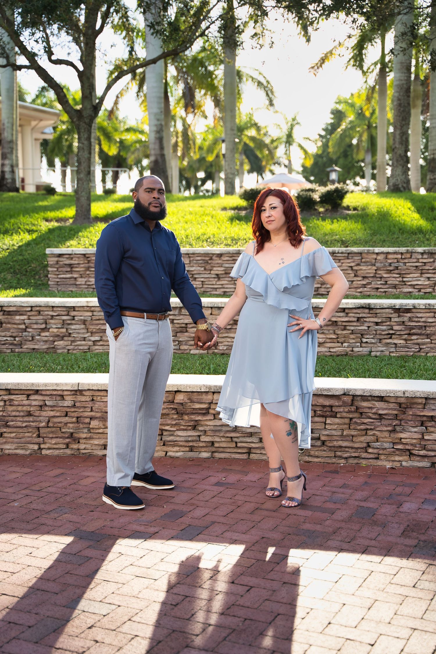 A stylish couple in coordinated blue and grey outfits pose together on brick pavement near palm trees.
