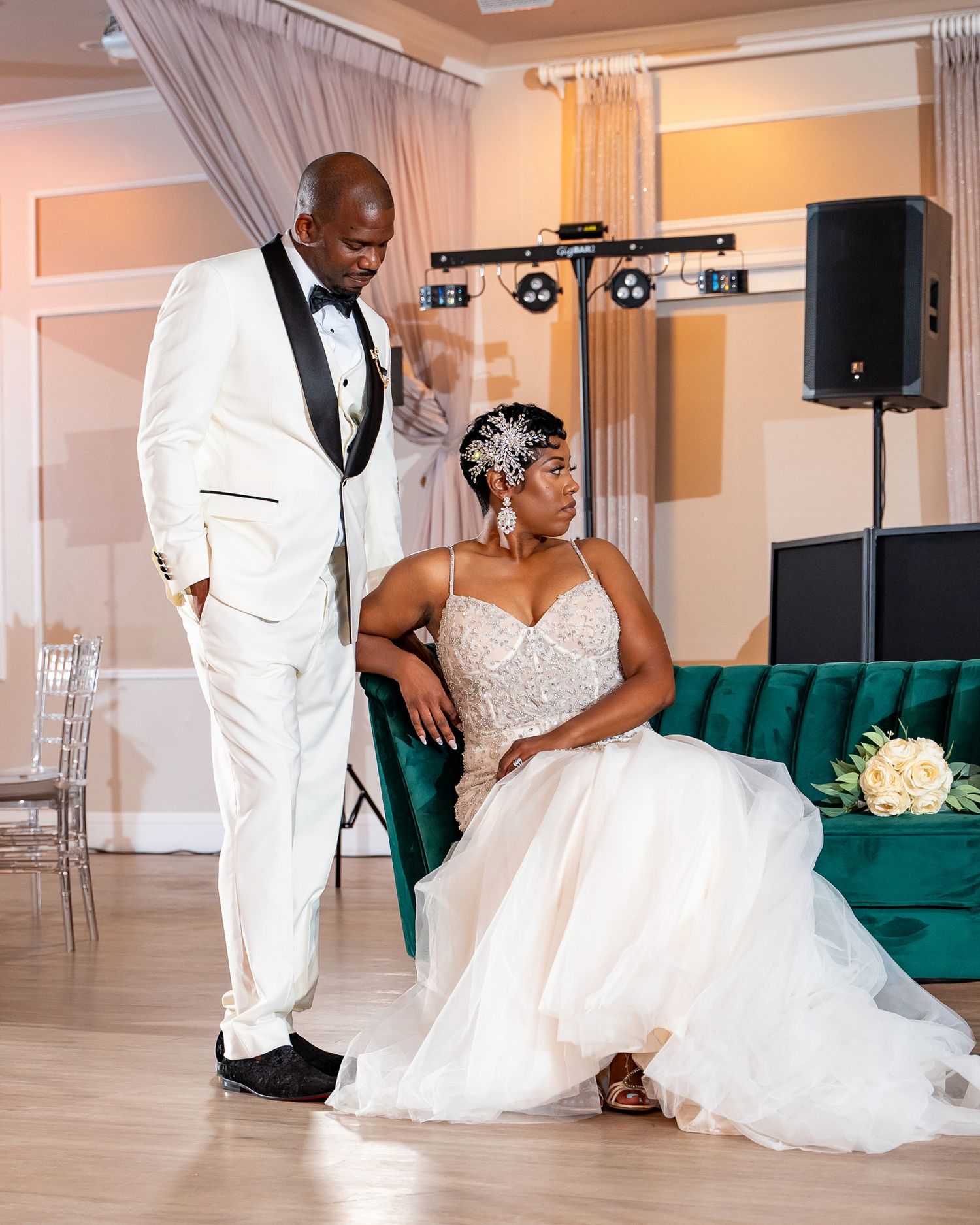 A formal wedding portrait featuring a white tuxedo and flowing wedding gown against an emerald velvet couch.