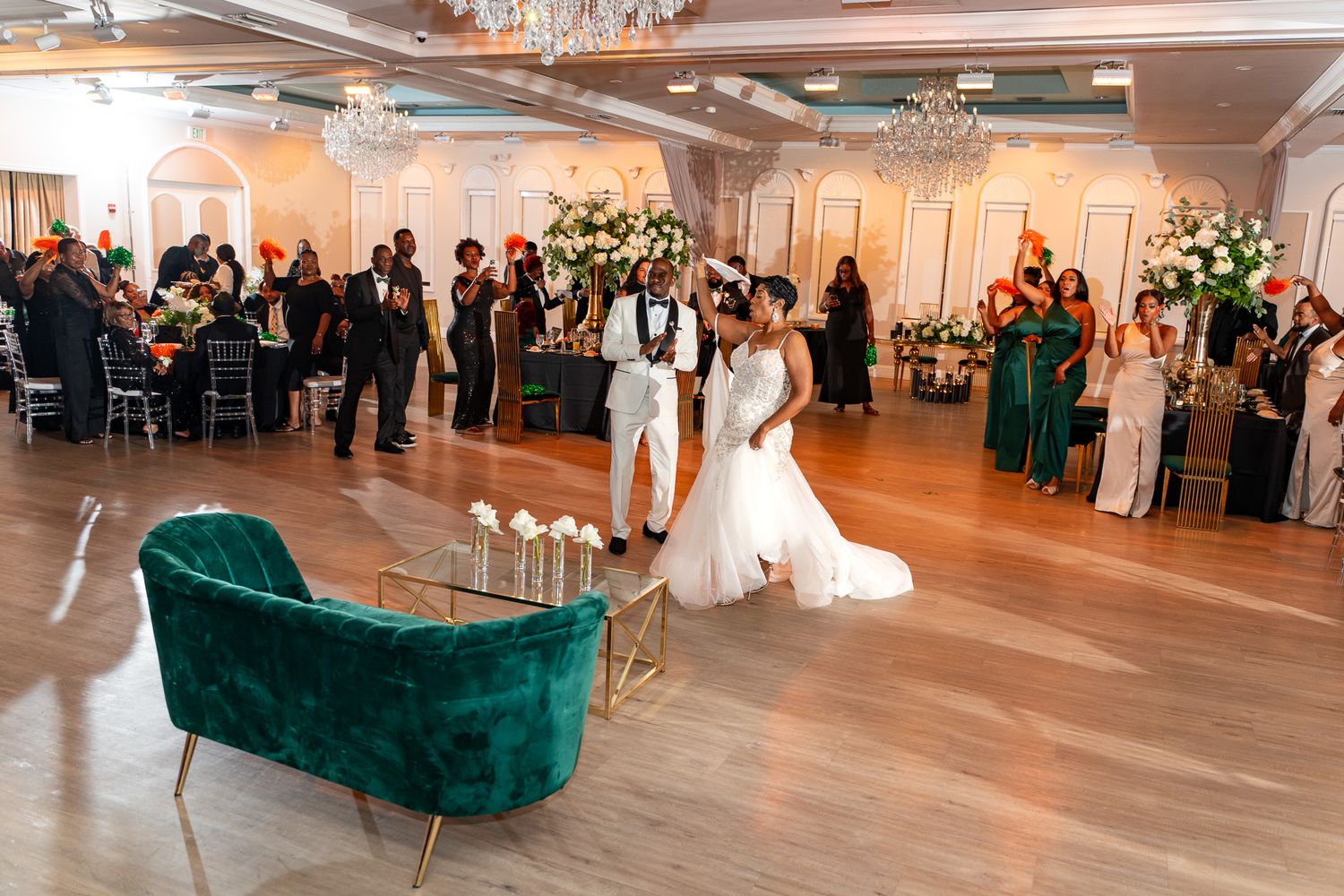 A couple performs their first dance in a luxurious ballroom decorated with chandeliers and white floral arrangements.
