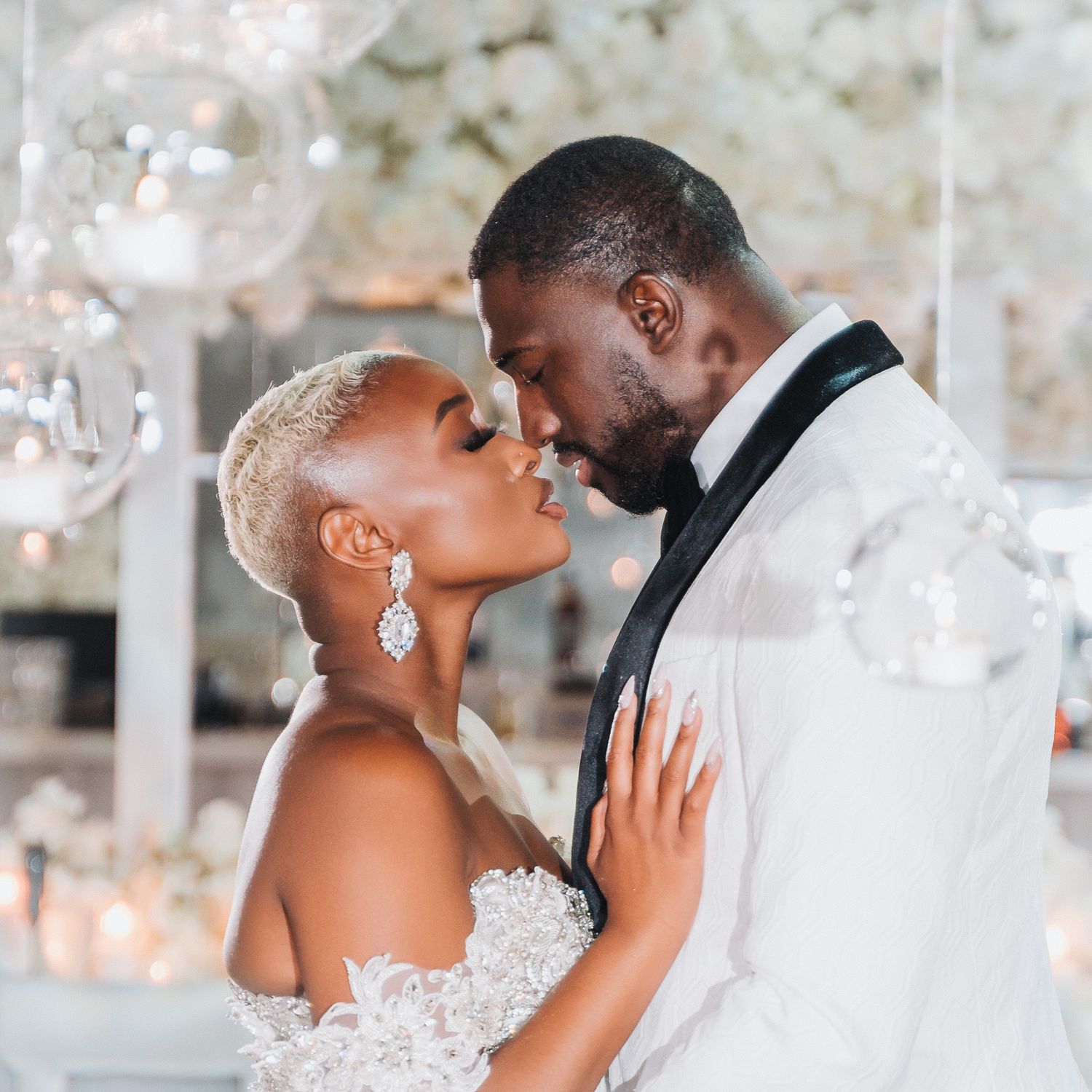 A romantic portrait of newlyweds in white attire sharing an intimate moment under crystal chandeliers.