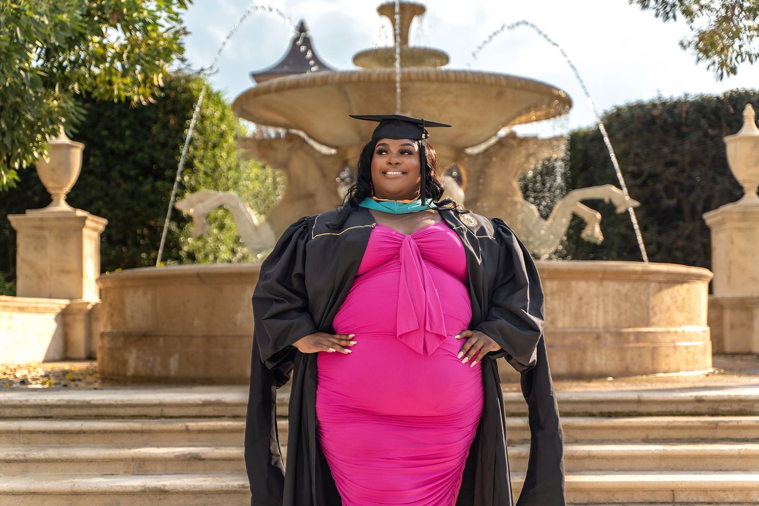 Graduate in pink dress and academic regalia poses by ornamental fountain during graduation.