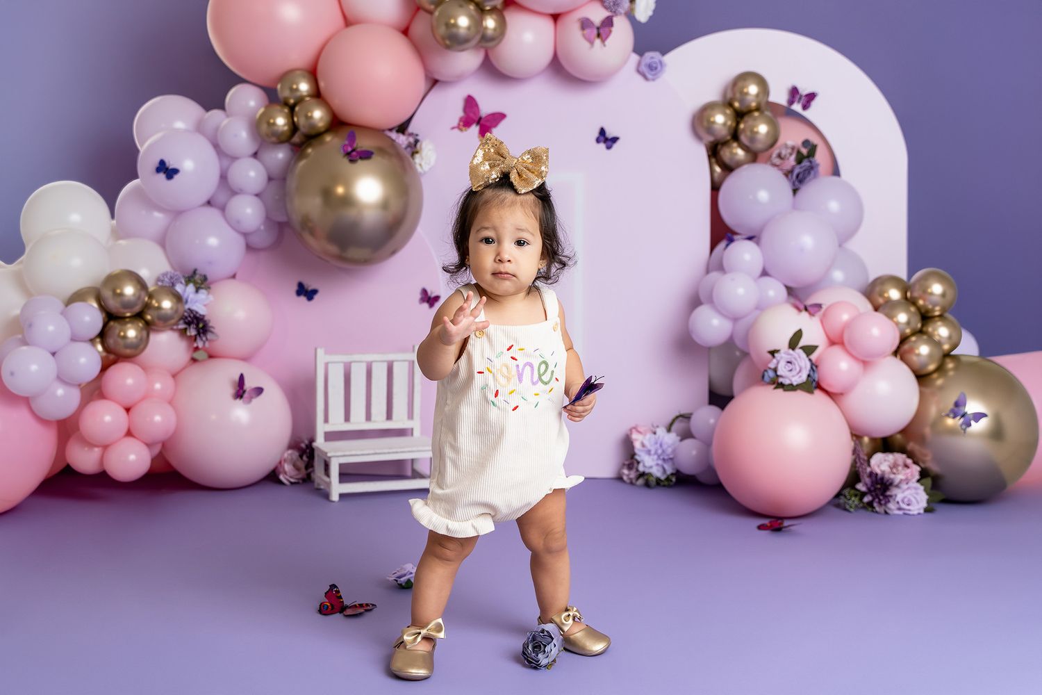 A birthday celebration setup with pink and purple balloon arch featuring a small white chair and butterflies.