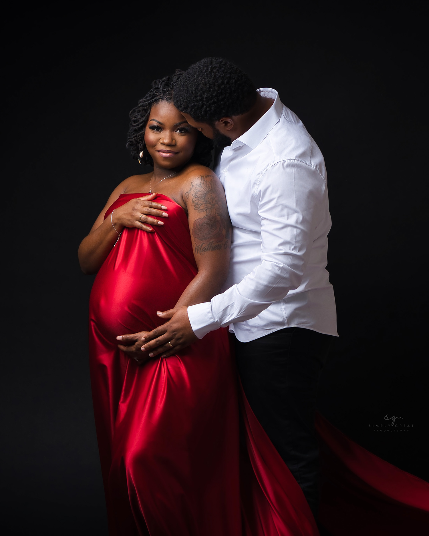 Intimate maternity portrait with couple in red dress and white shirt against black background.