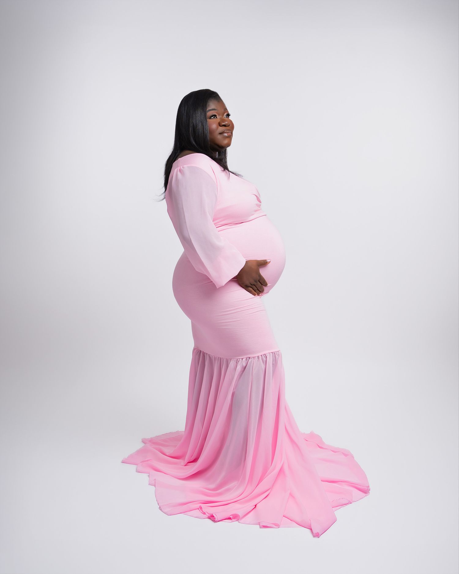 Side view of pregnant woman wearing flowing pink maternity gown in studio setting.