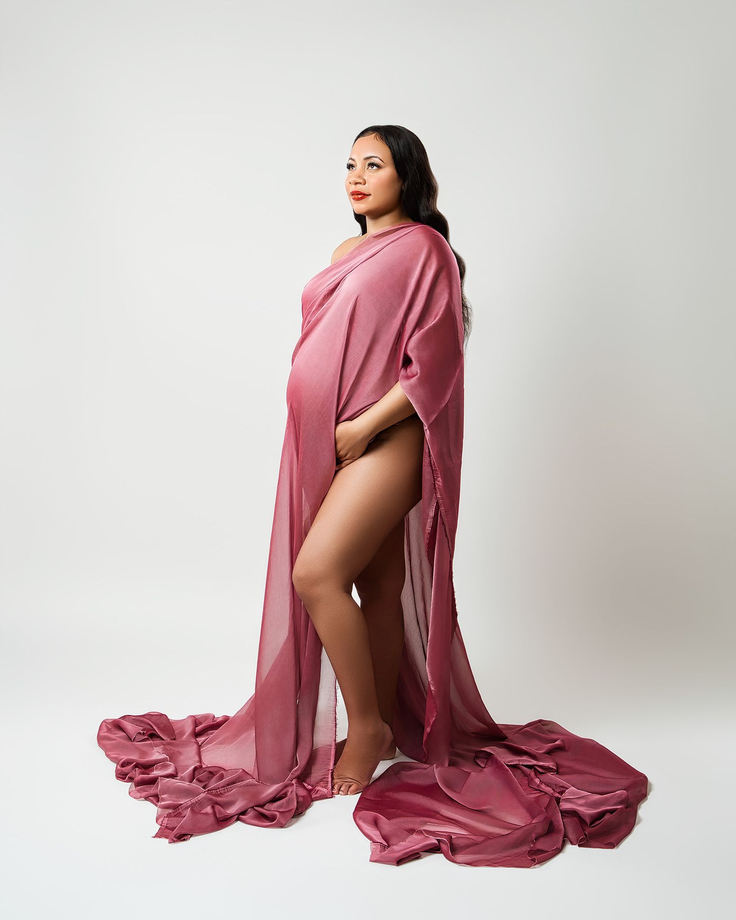 Dramatic maternity photo with flowing rose-colored fabric against white backdrop.