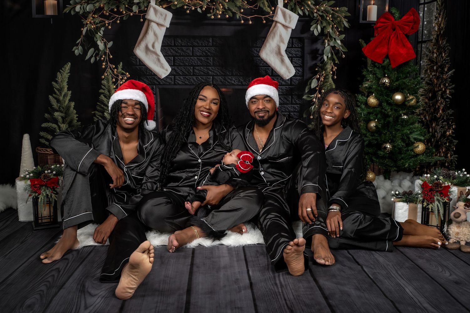 Group of people wearing holiday attire pose together on wooden floor with Christmas decorations in background.