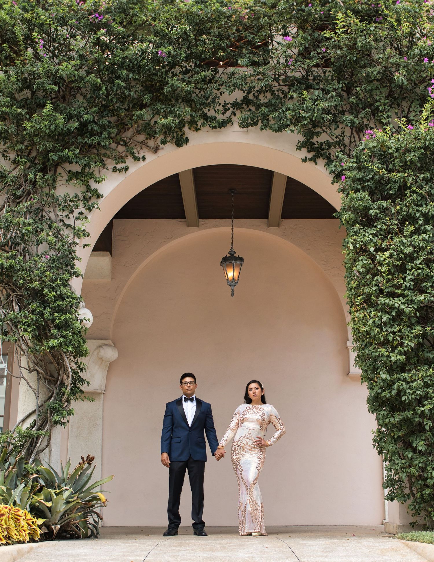 Wedding couple stands under Mediterranean archway surrounded by lush greenery.