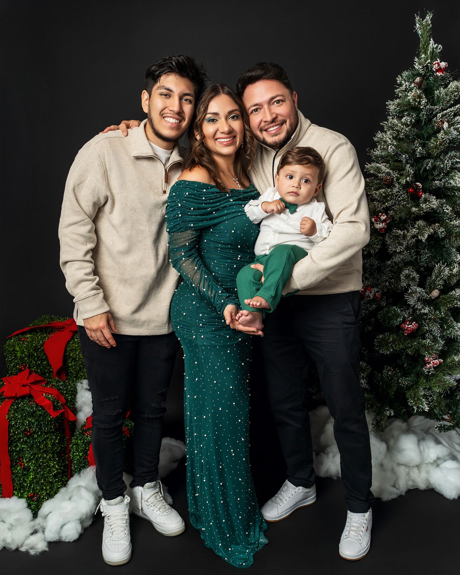 A family poses in matching cream sweaters by a Christmas tree against a dark backdrop.