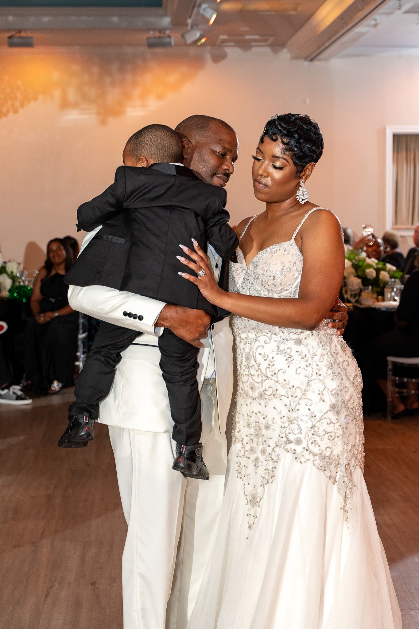 Sweet moment as groom in white suit holds a small child while dancing with bride at wedding reception.