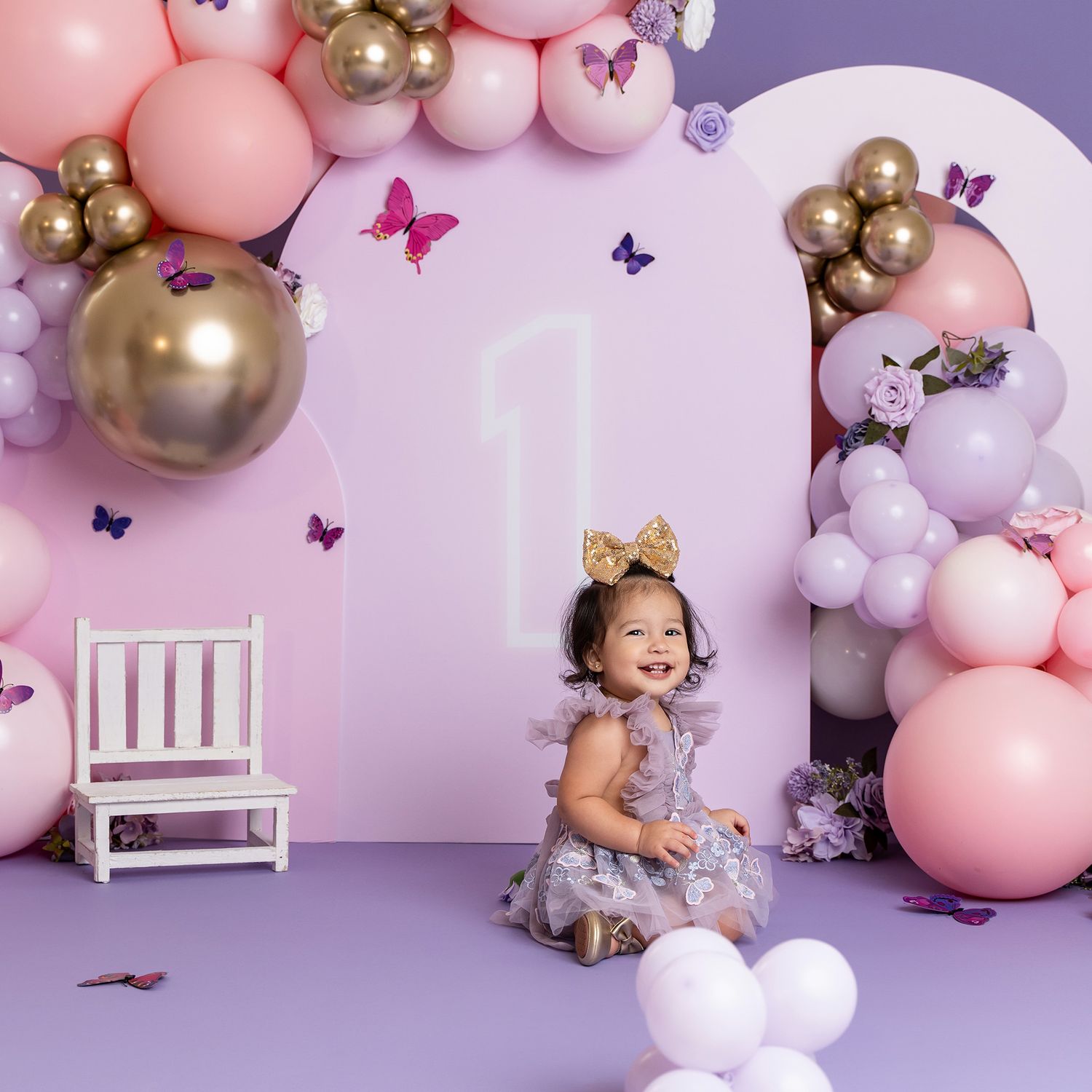 Birthday photoshoot setup with pink and gold balloons, white chair, and purple butterfly decorations.