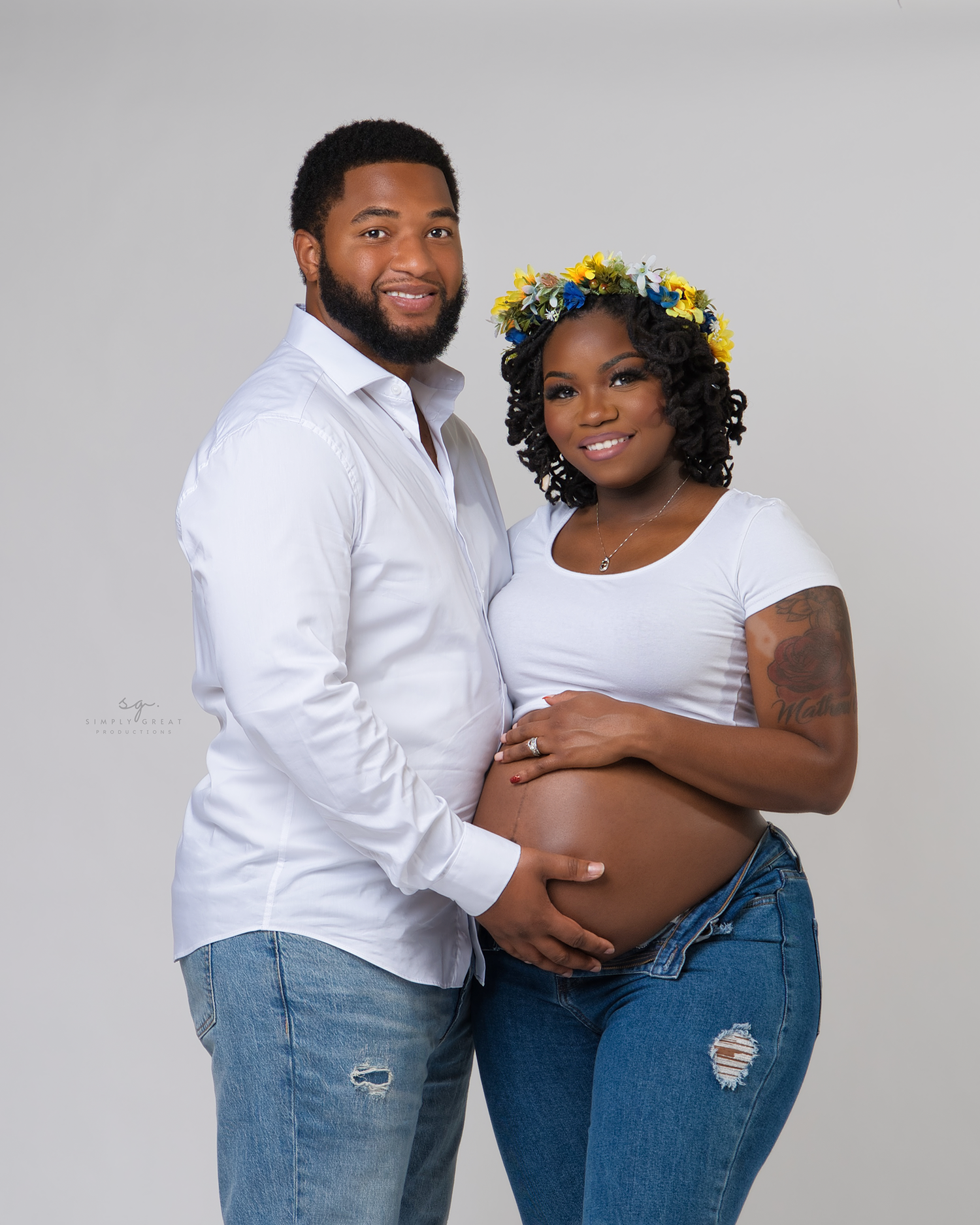 Studio maternity portrait of couple in matching white tops and distressed jeans.