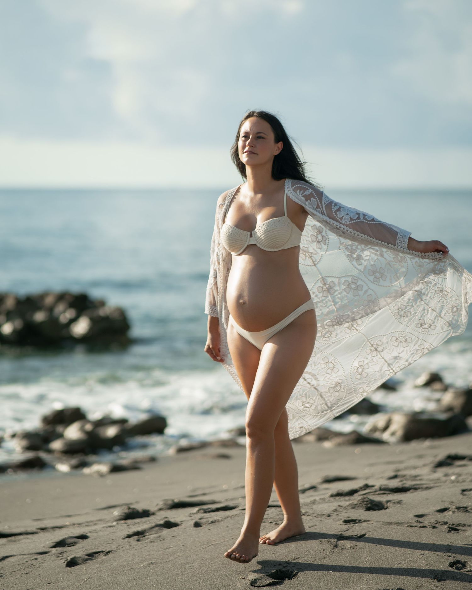 Expectant mother in white beachwear and flowing lace robe poses gracefully on rocky shoreline.