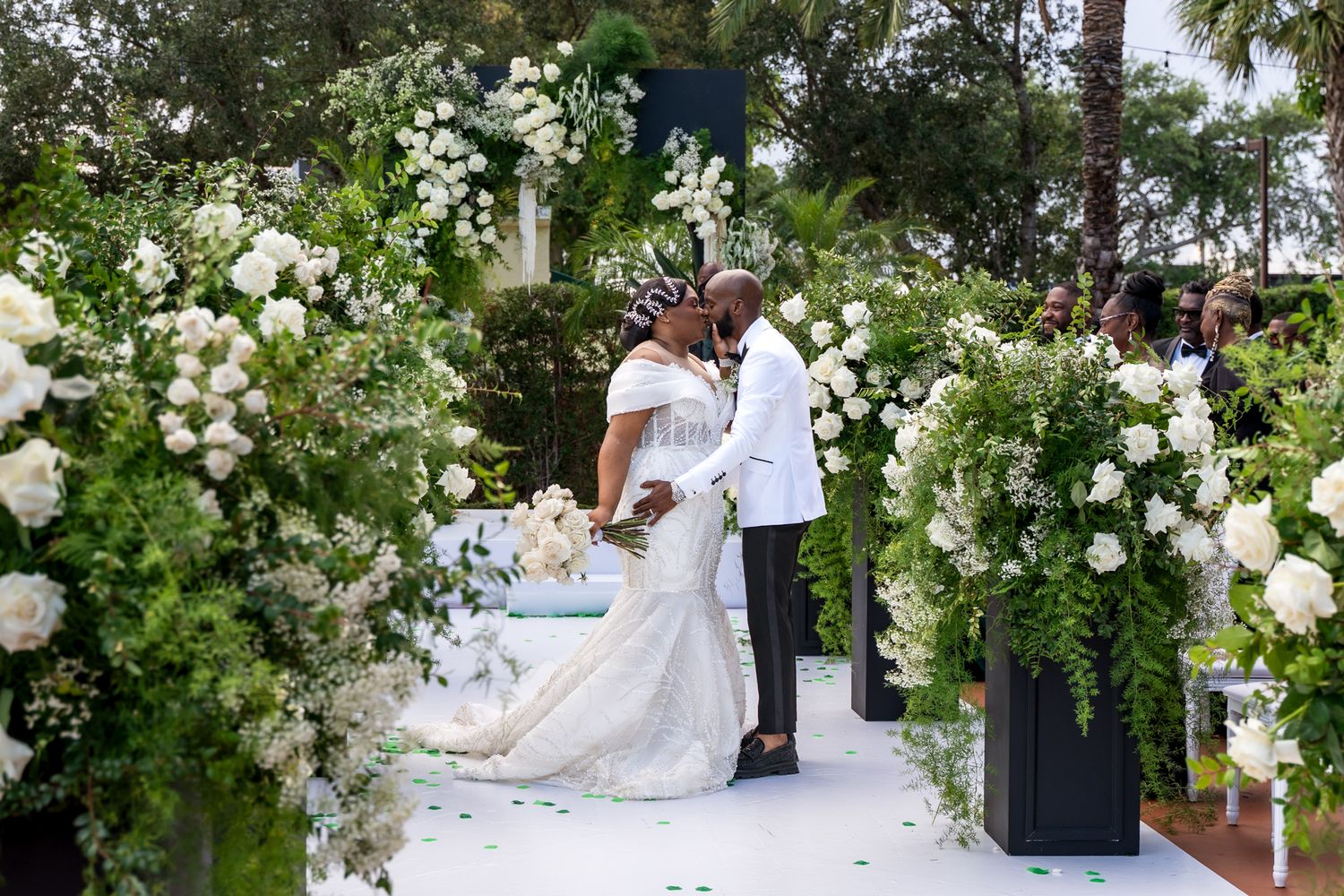 A couple shares a tender moment under an elegant white floral arch during their outdoor garden wedding ceremony.