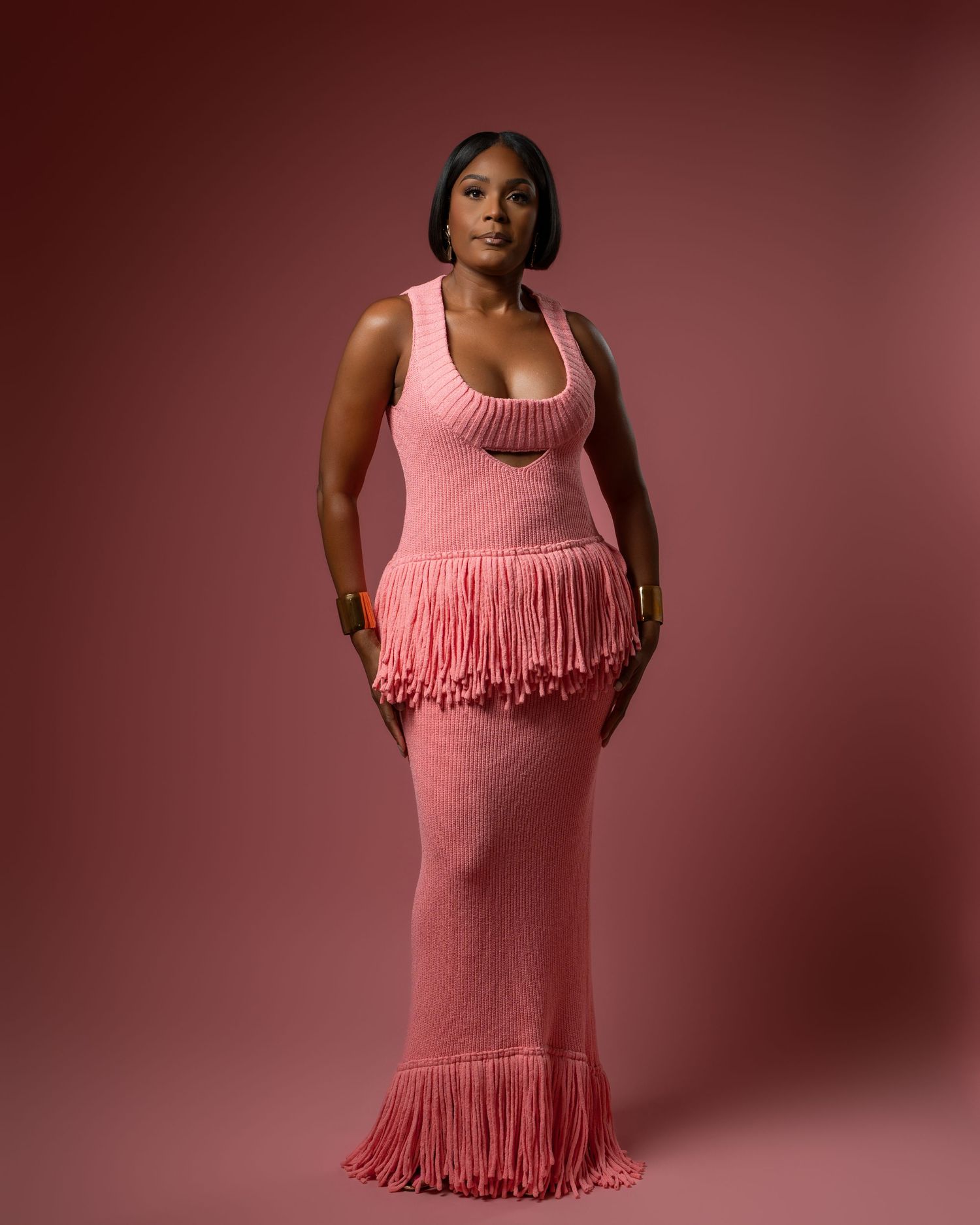 A model in a pink knit dress with fringe detail poses against a burgundy background in a fashion photoshoot.