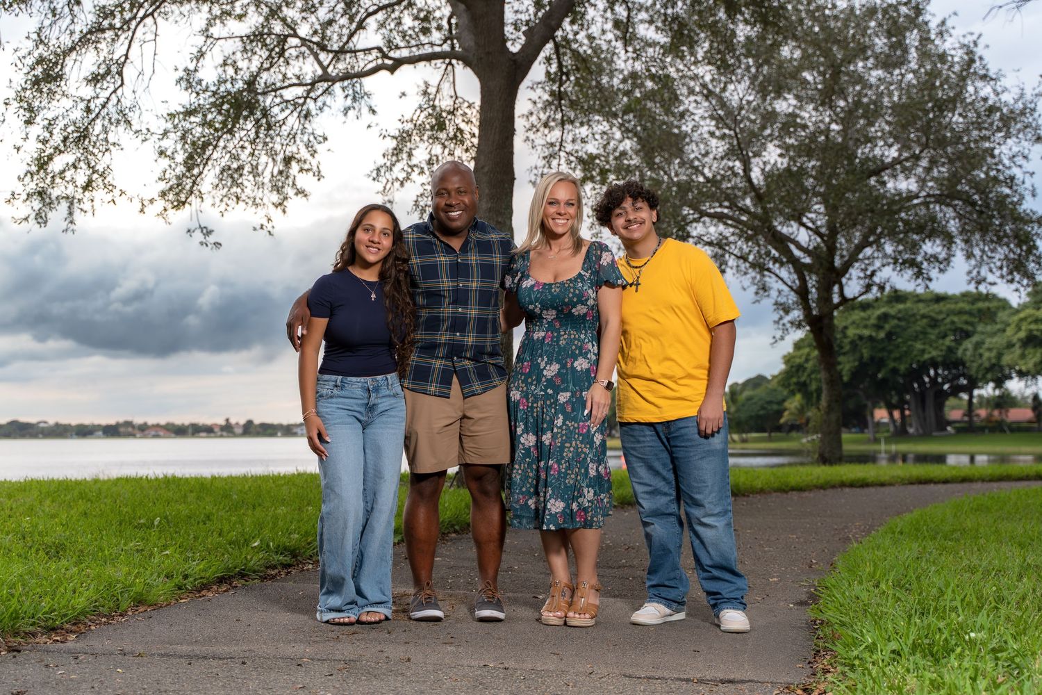 A group of people standing together on a path near a lake on a cloudy day with trees in the background.