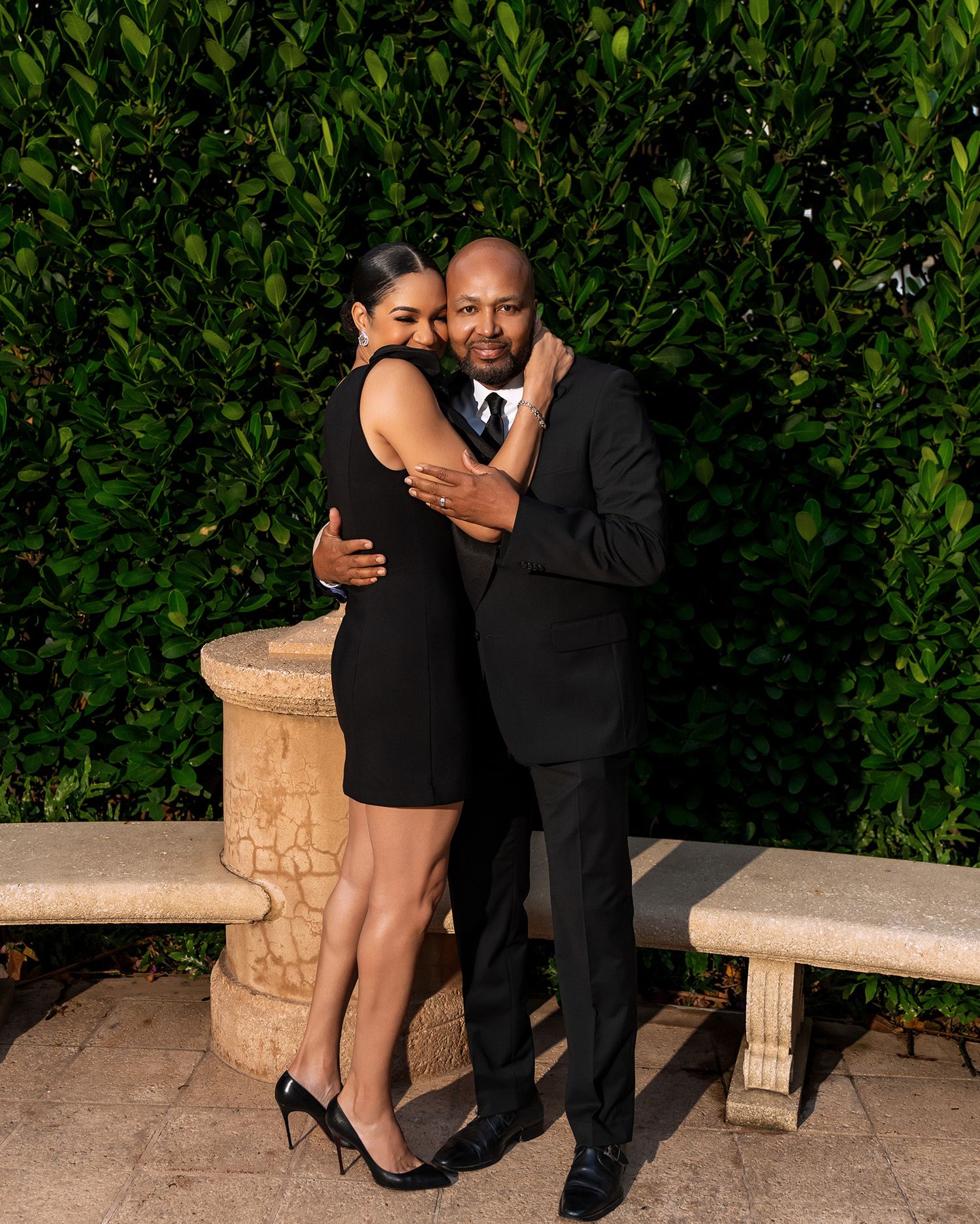 A couple embraces while standing on stone benches against a green hedge wall.