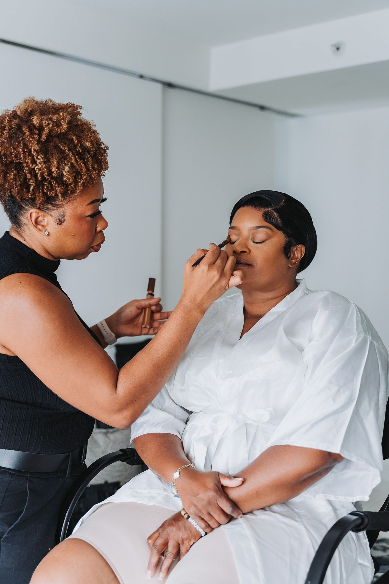 A makeup artist applies cosmetics to a client wearing a white robe during wedding preparation.