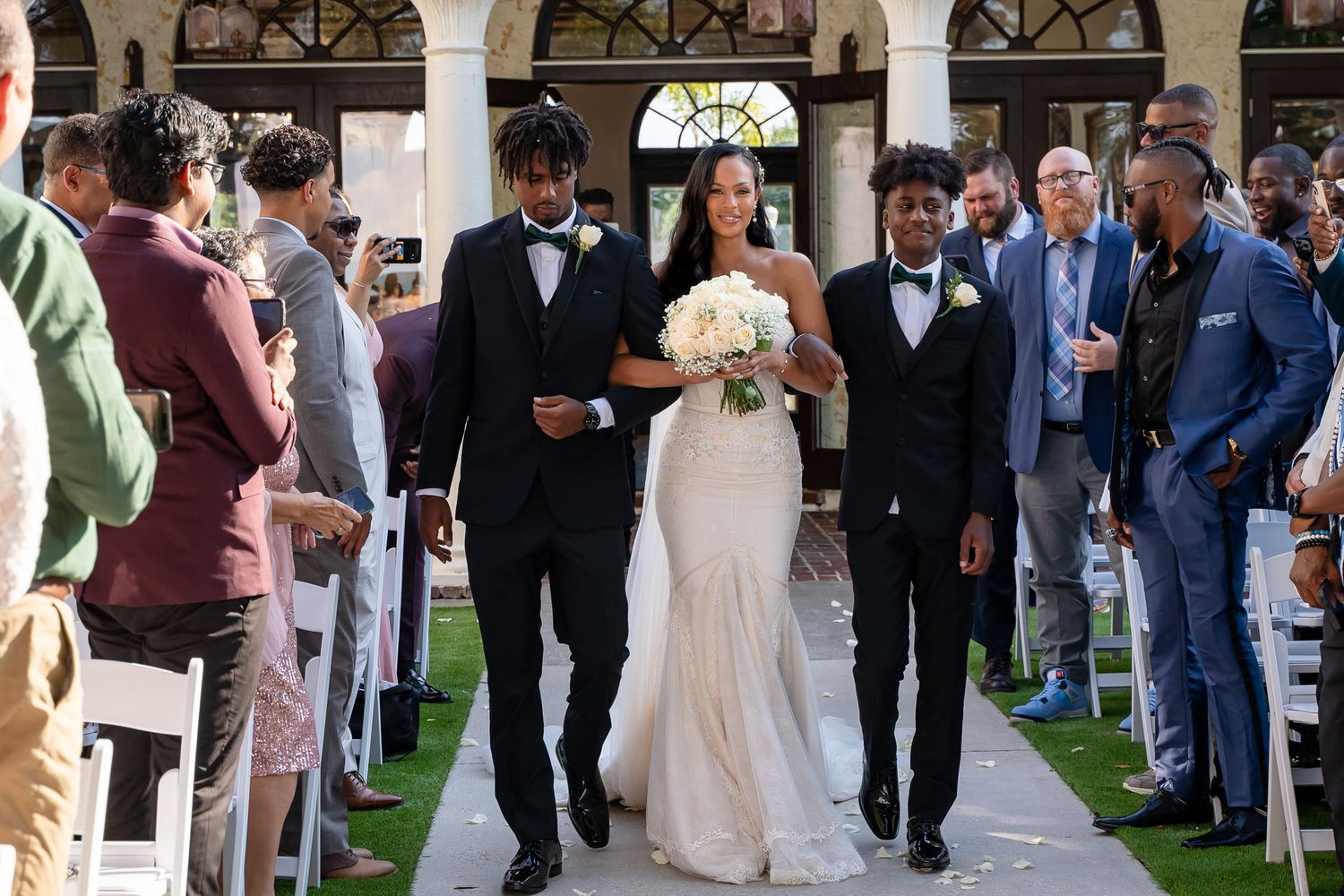 A wedding party walks down an aisle lined with guests under a columned pavilion.
