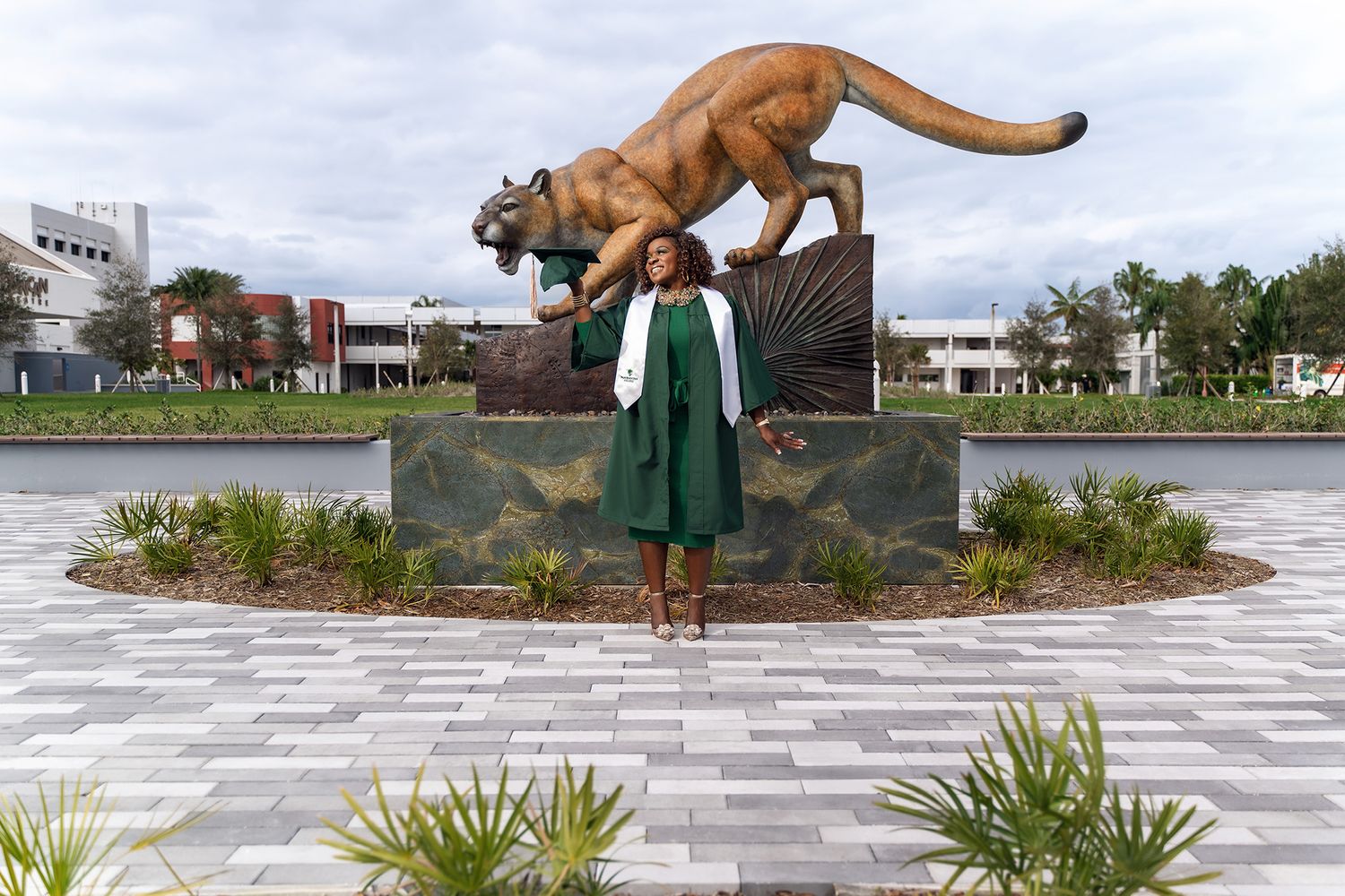 Graduate in green dress and white stole poses beneath large bronze mountain lion statue.