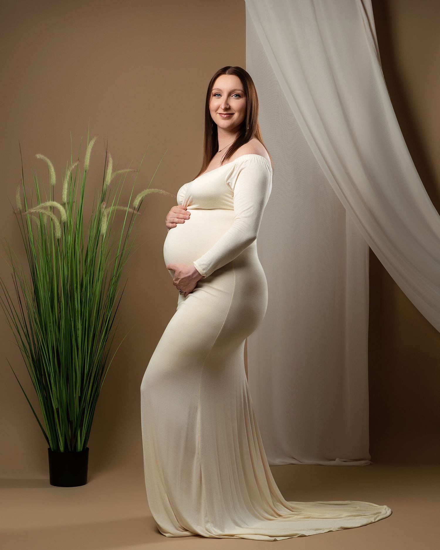 Elegant maternity portrait with woman in cream dress posing beside decorative plants and drapes.