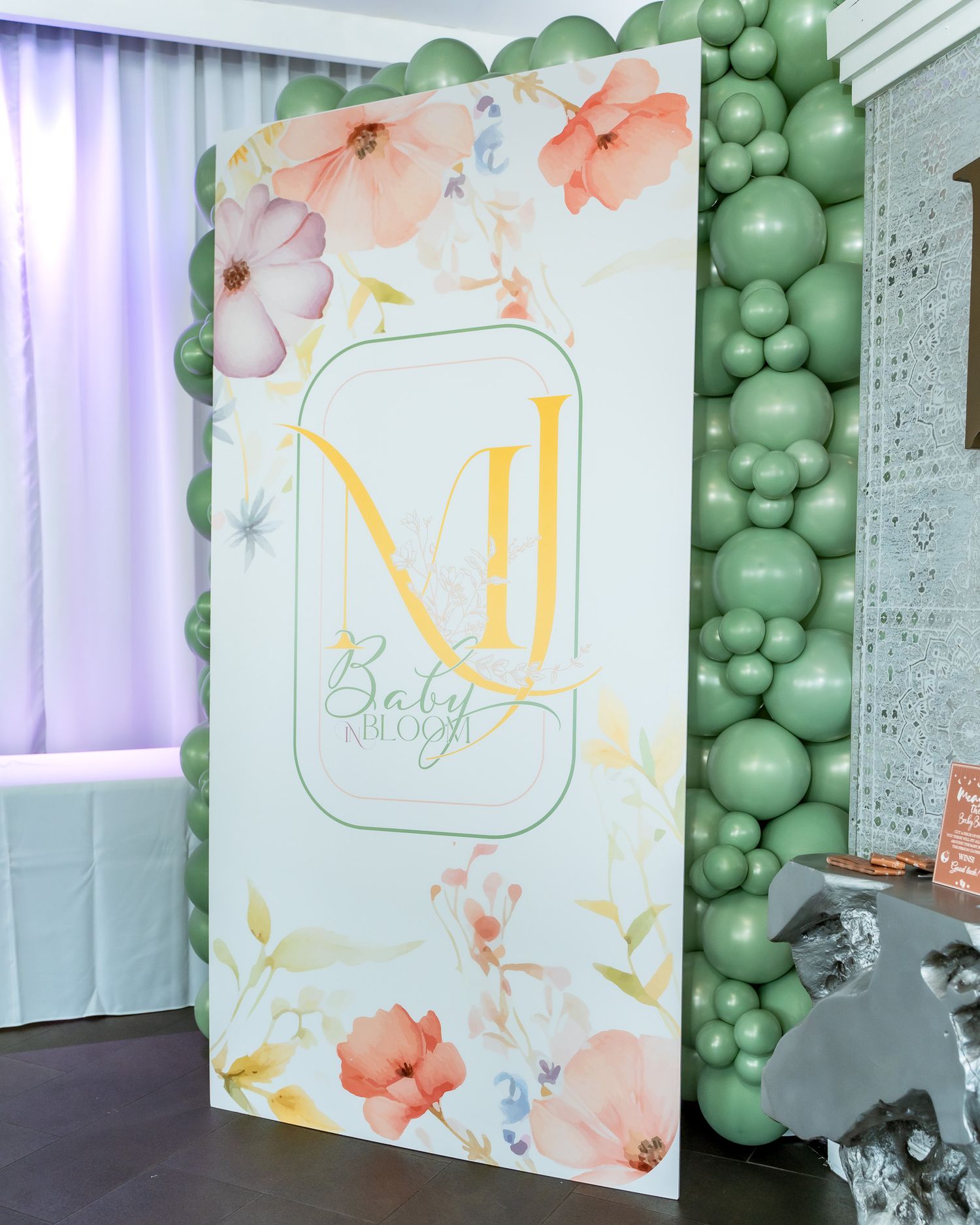 Decorative baby shower backdrop with floral design, green balloon arch and monogram logo.