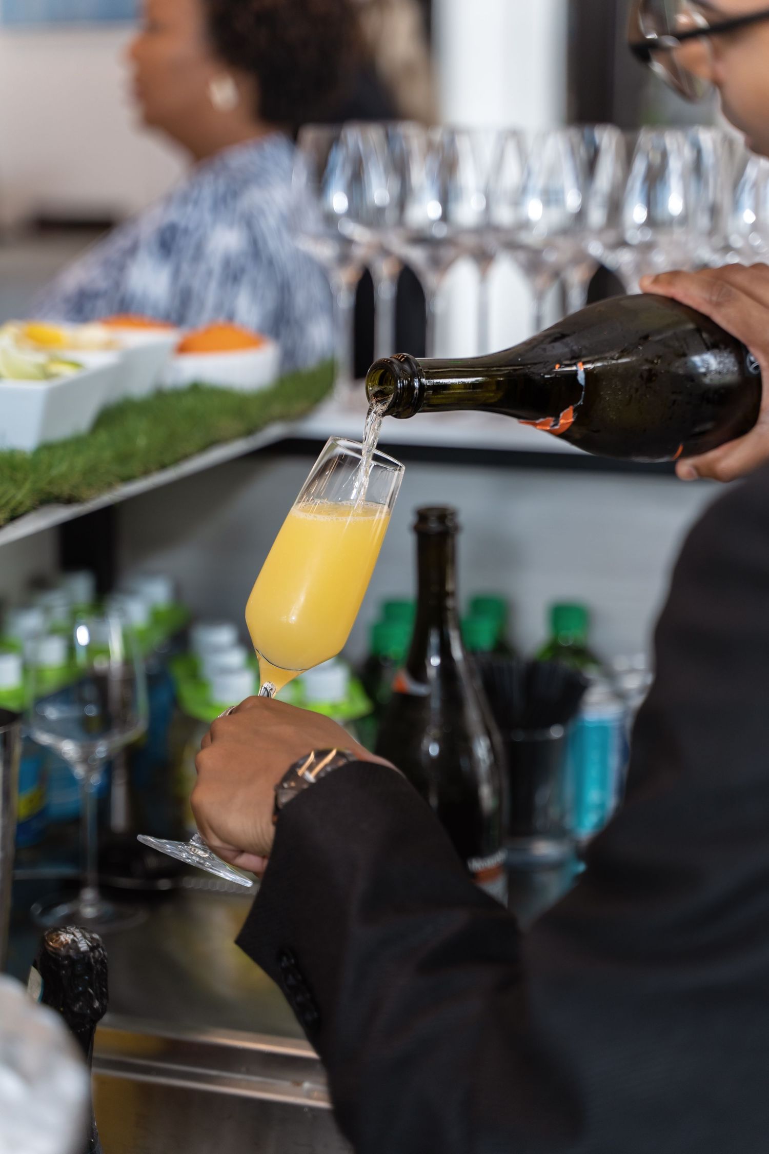 A bartender pours orange juice from a dark bottle into a champagne flute at an elegant bar setting.
