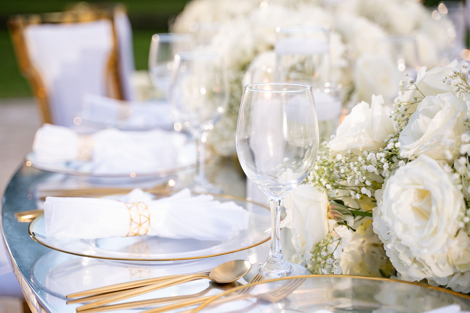 Elegant wedding table setting with white roses, crystal glasses and gold cutlery at an outdoor reception.