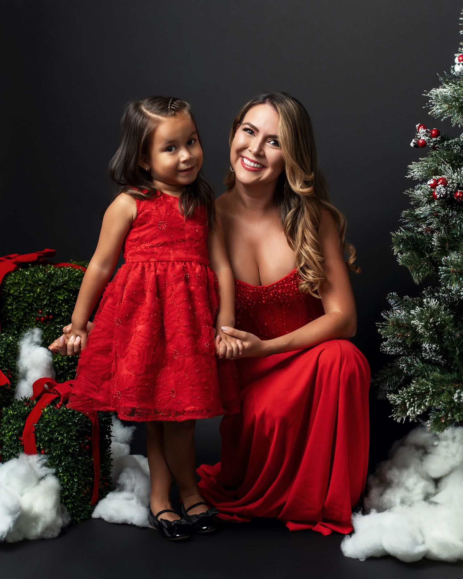 A festive holiday portrait with matching red dresses against a Christmas tree backdrop with cotton snow on the floor.
