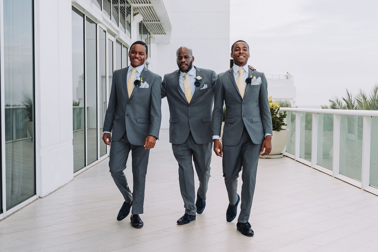 Three groomsmen in matching gray suits walk confidently down a modern hallway with glass windows.