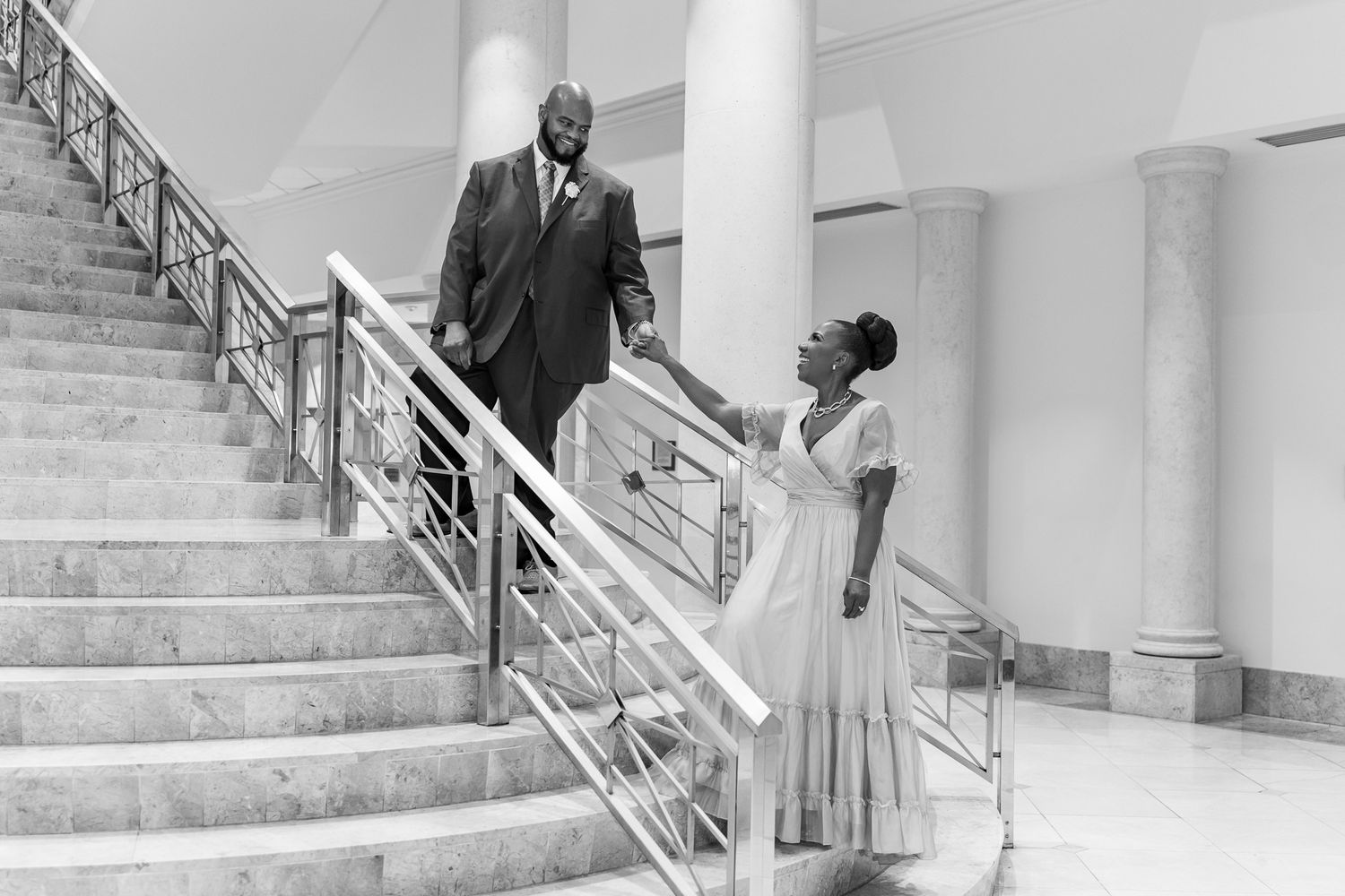A black and white photograph of a couple descending an elegant staircase with white columns.