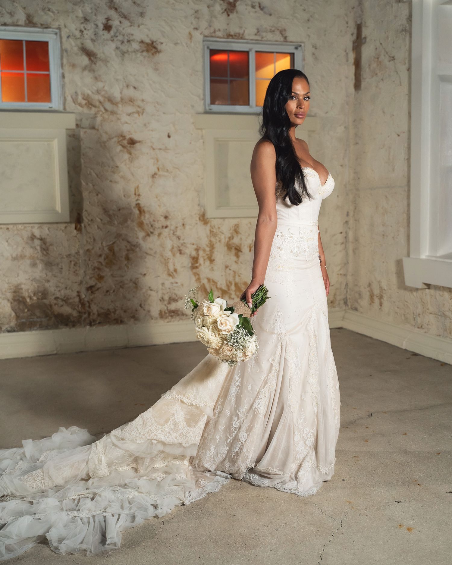 A bride in a flowing white lace gown holds a bouquet in an industrial-style setting.