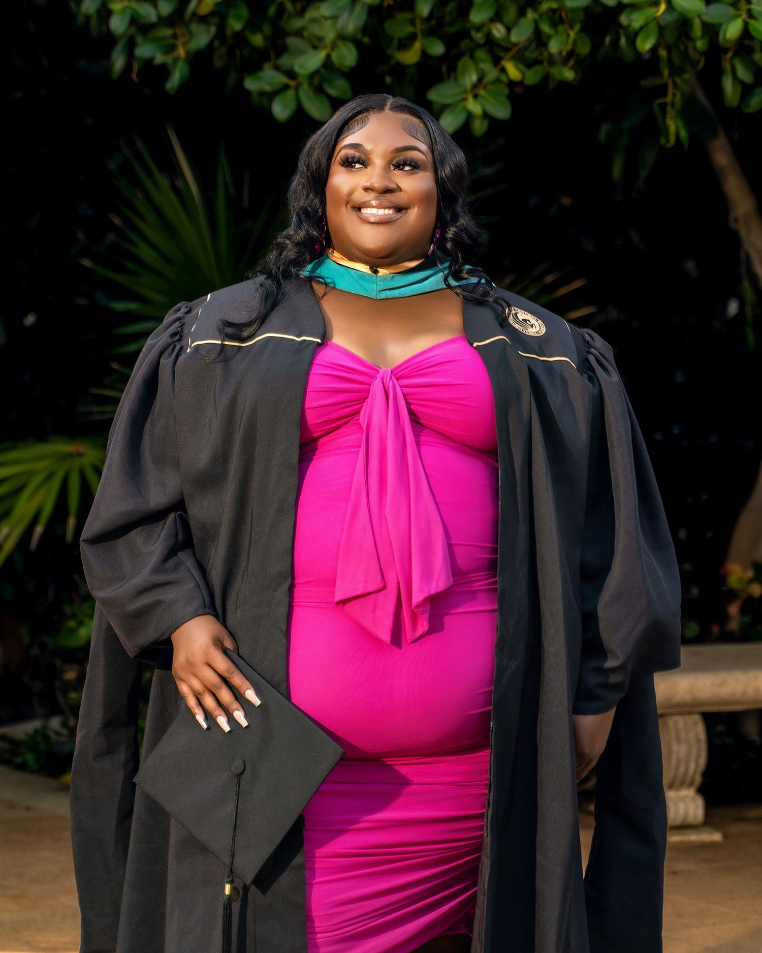 Graduate in pink dress and academic gown poses with diploma outdoors.