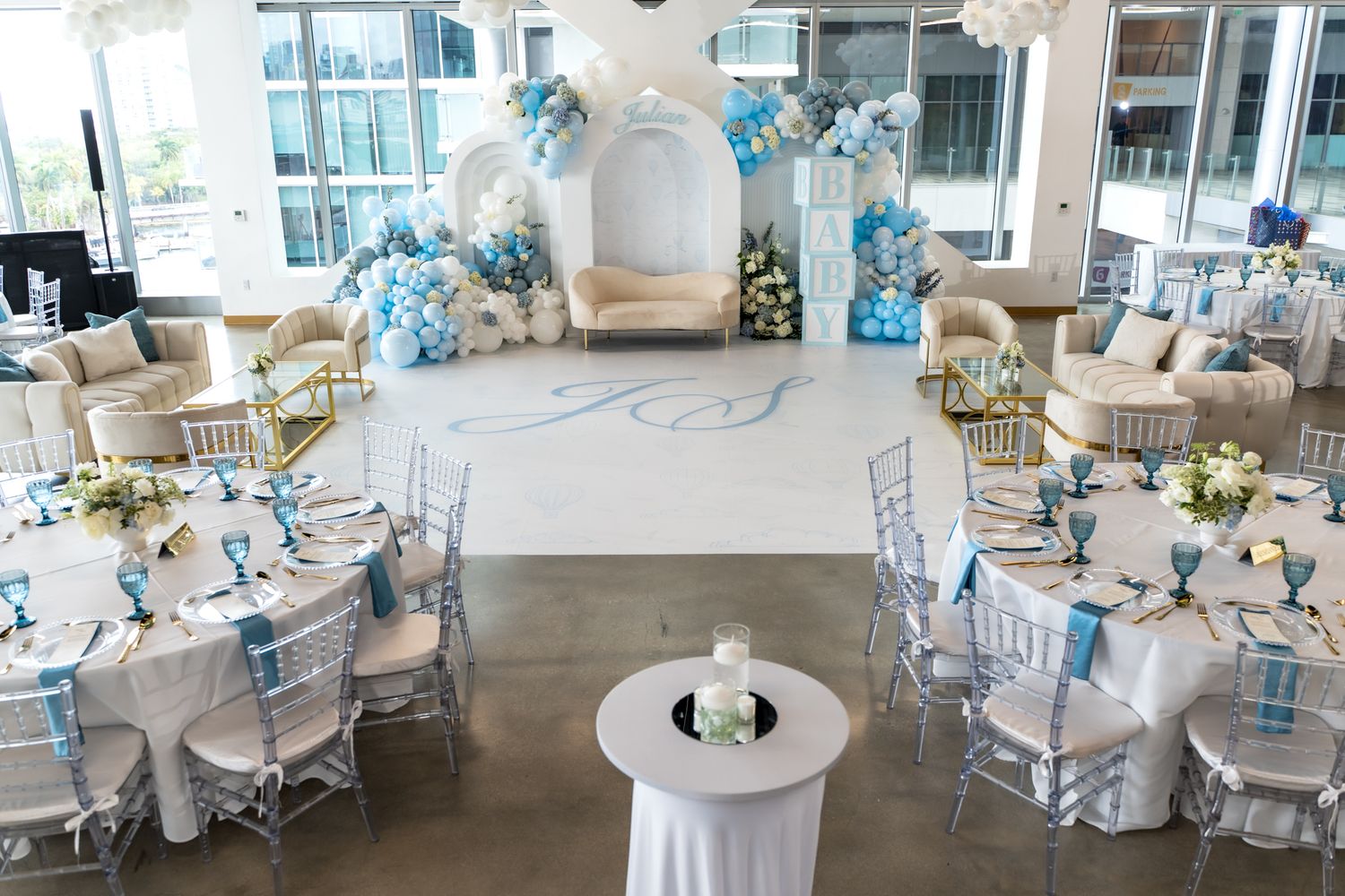 Elegant baby shower setup with white and blue decor featuring round tables and a ceremonial arch.