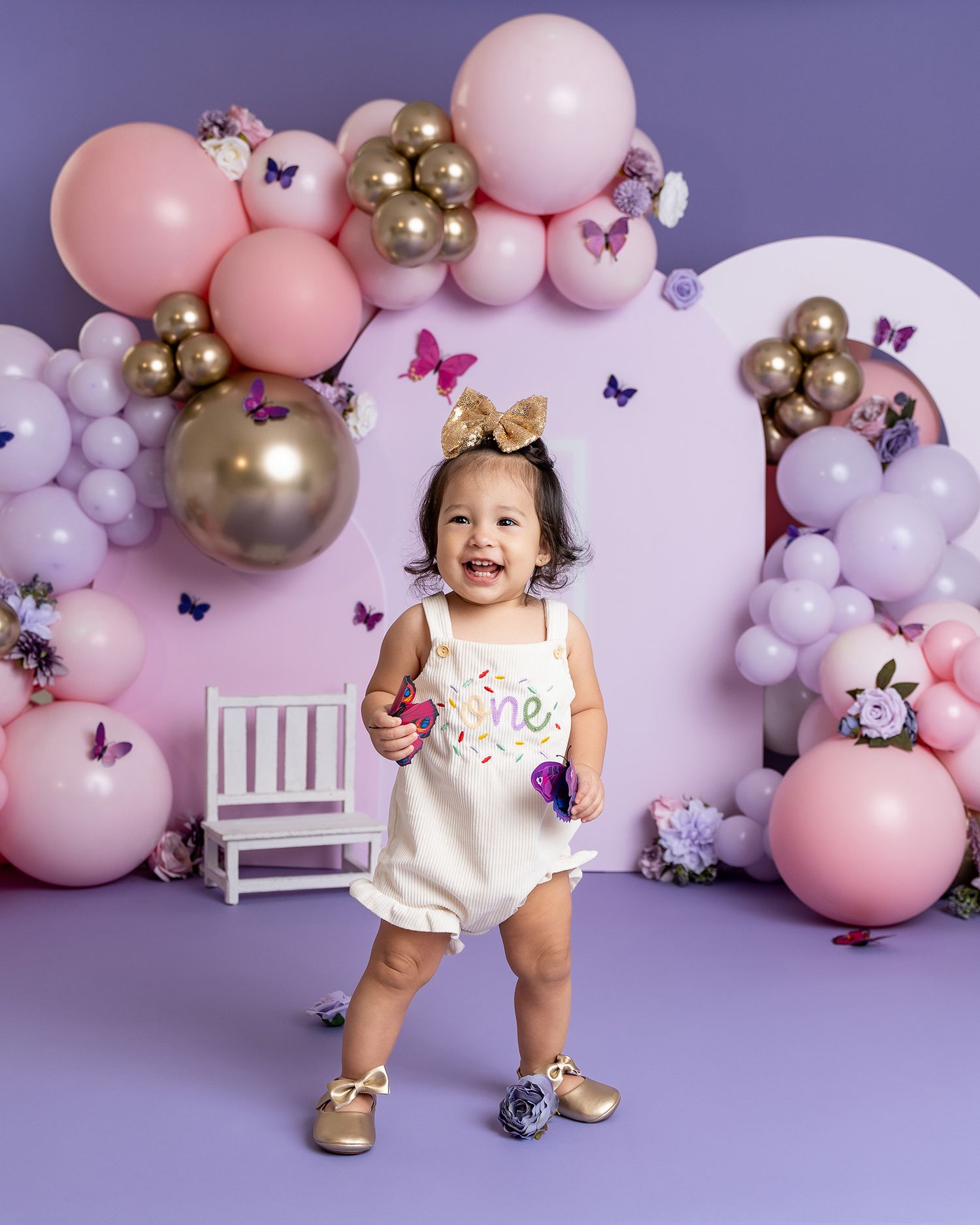 A toddler in a white outfit poses happily among pink and gold balloons with butterfly decorations.