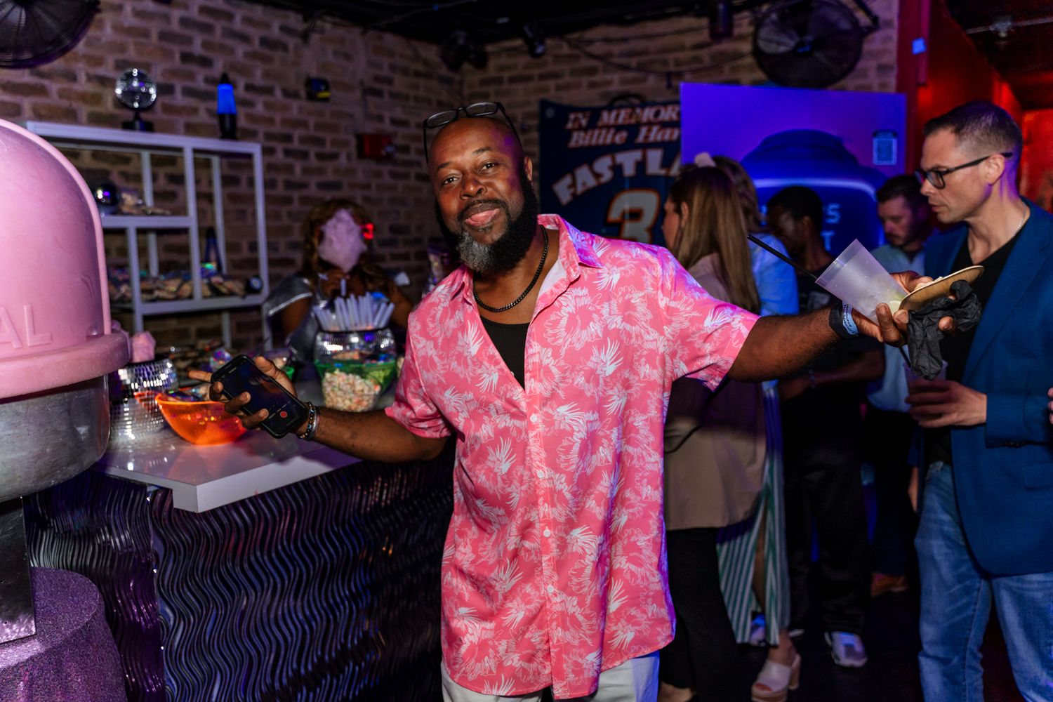 Man in pink Hawaiian shirt smiling at bar counter during nighttime event.