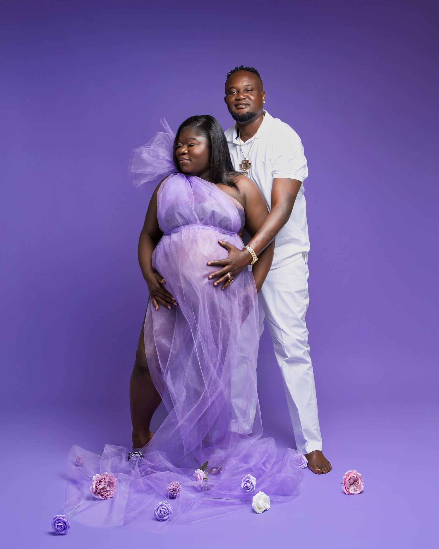 Couple's maternity portrait with purple tulle gown against purple background with scattered roses.