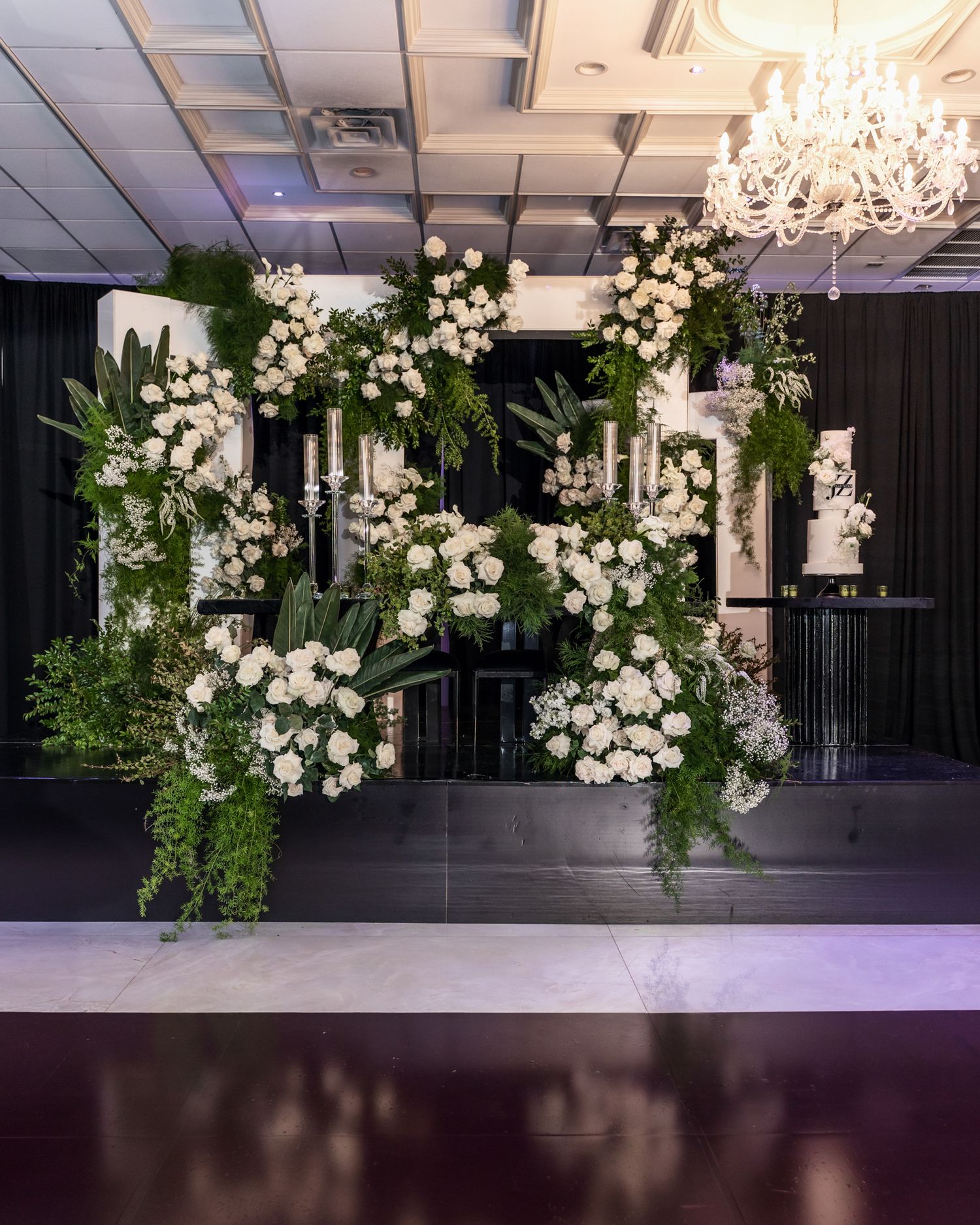 Indoor wedding venue decorated with cascading white flowers and greenery against dark walls and crystal chandeliers.