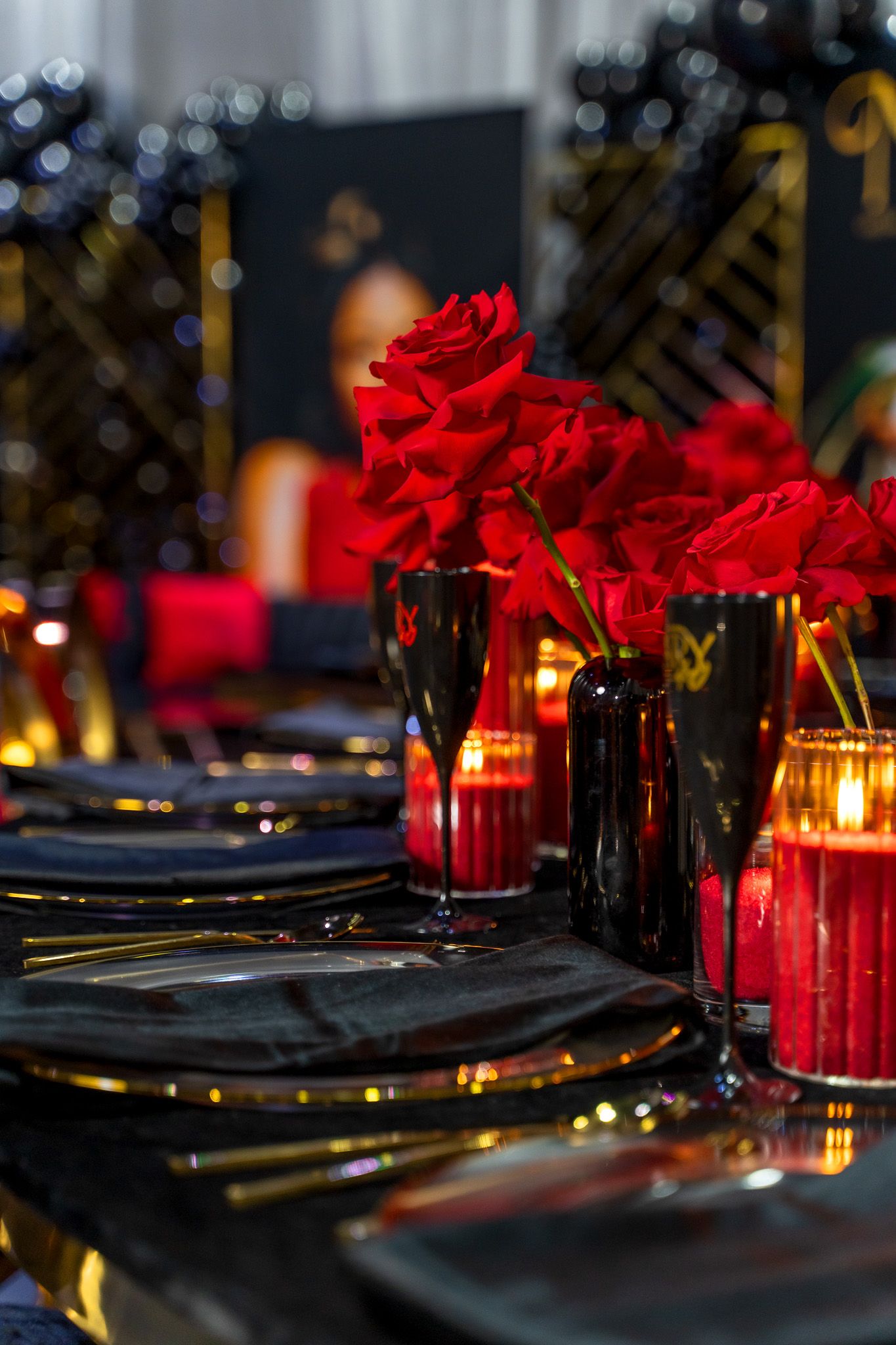 Elegant table setting with red roses and glowing candles in black vases against a dark background.