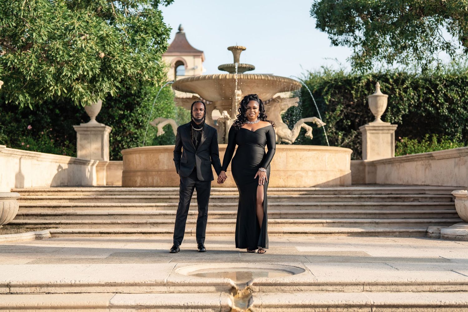 A couple in formal attire poses on elegant stone steps with ornate urns and fountain at an upscale outdoor venue.