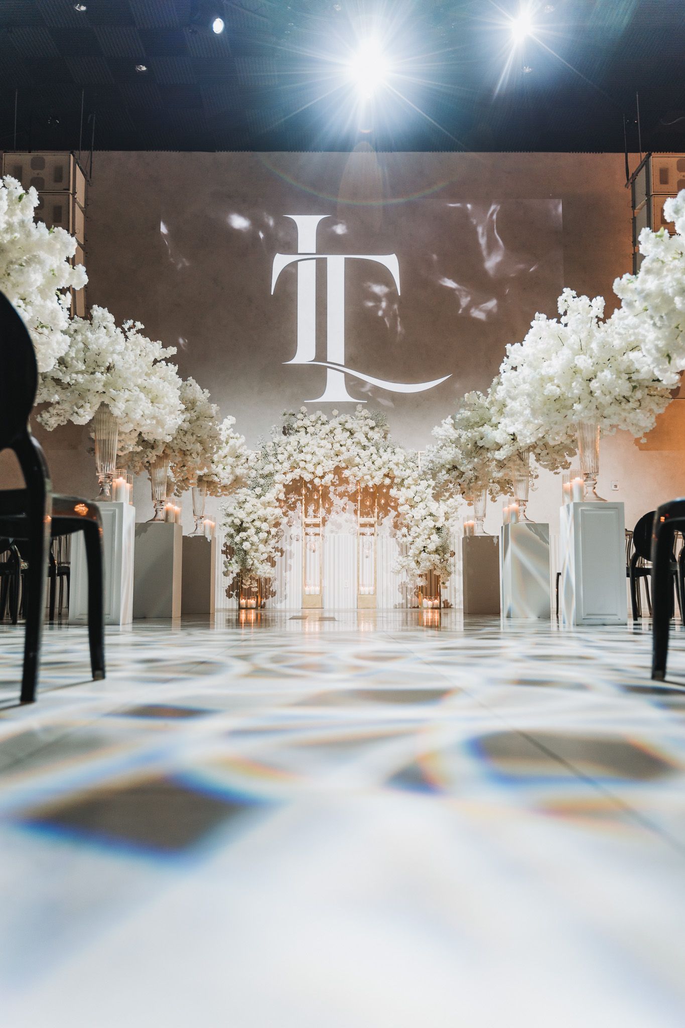 Elegant wedding venue setup with white floral arches and monogram projection on checkered floor.