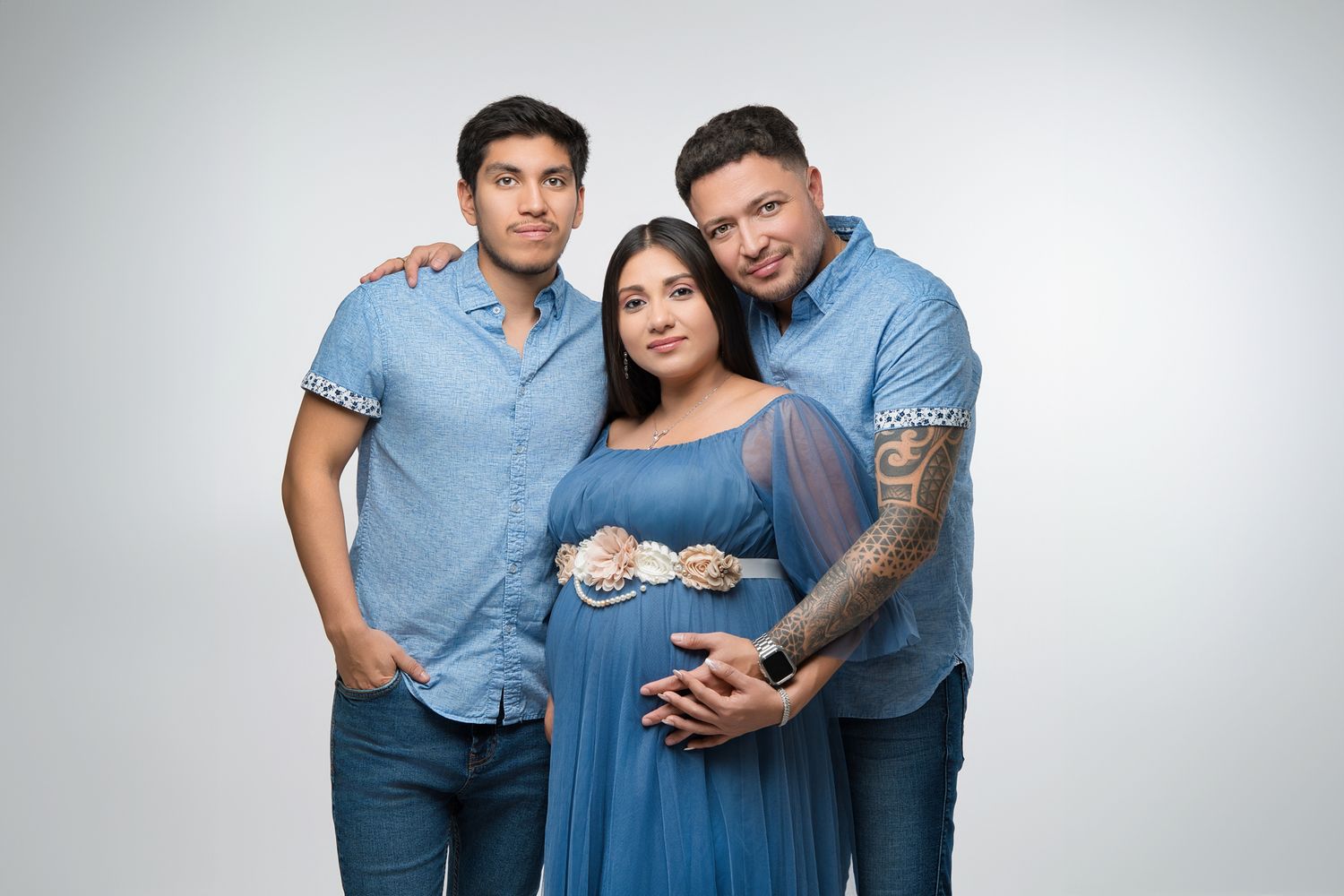 A maternity shoot features a trio in coordinating blue outfits posing together against a light background.