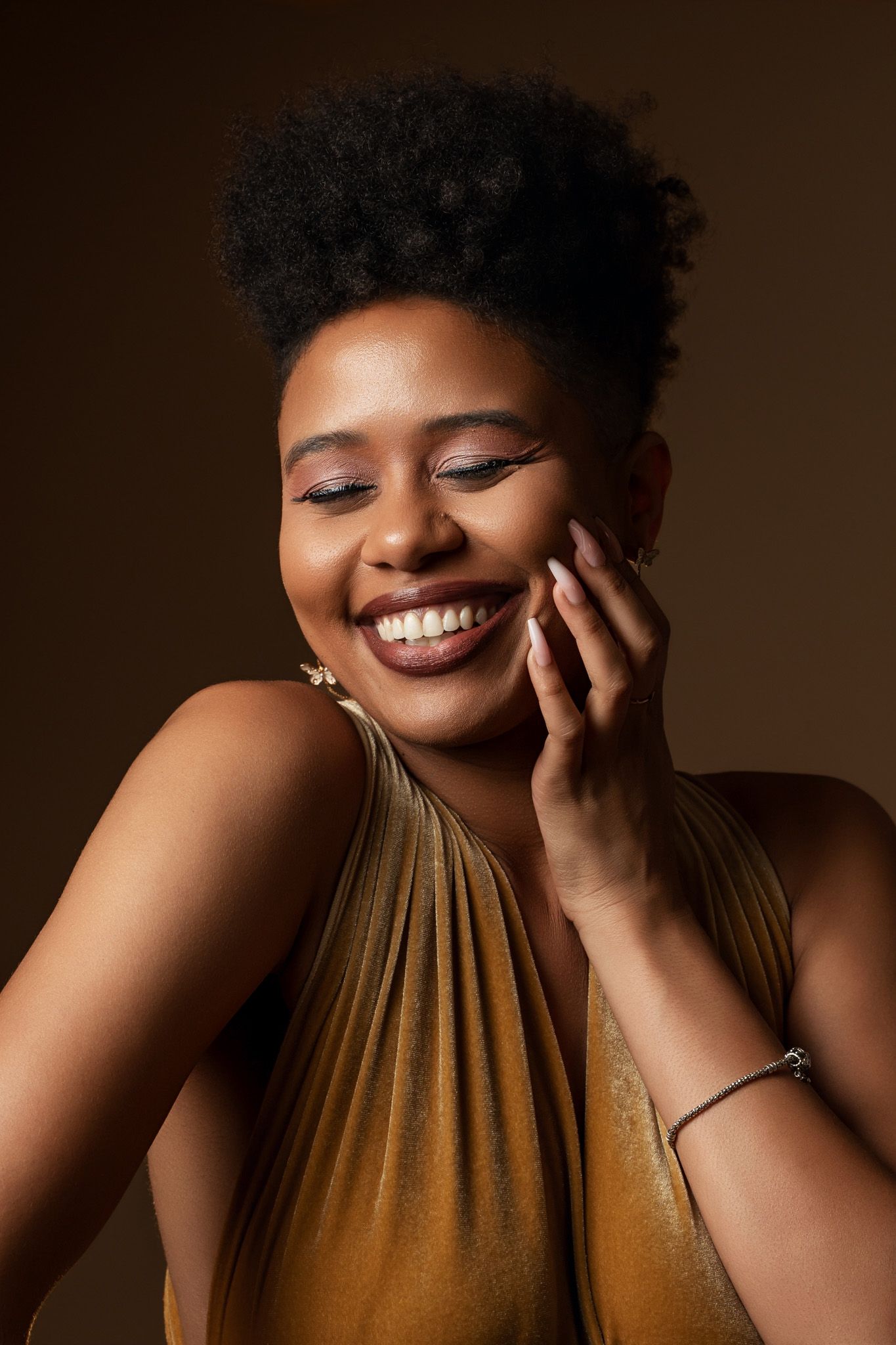 Close-up portrait showcasing joyful expression in gold halter dress against warm brown background.