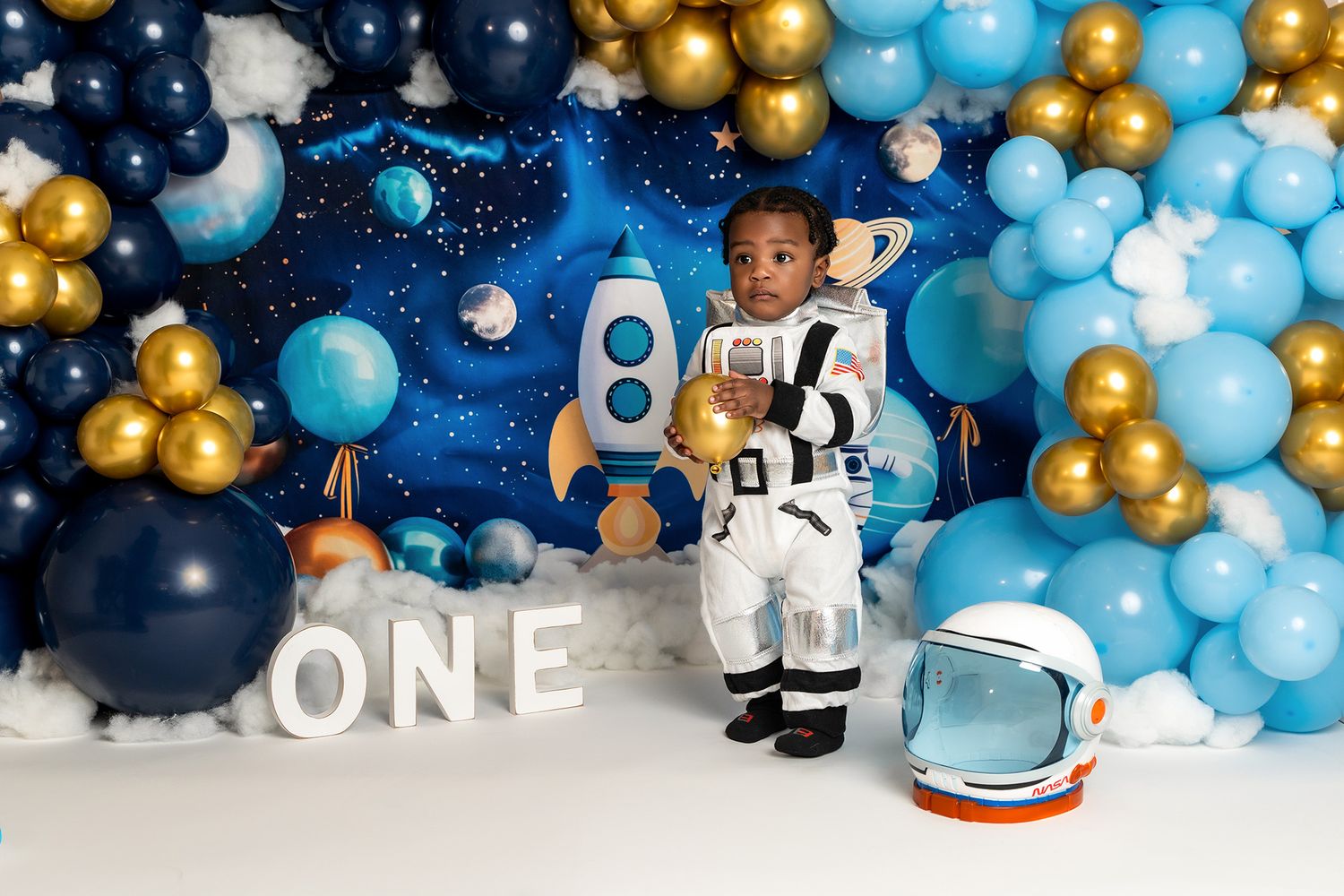 A one-year-old photoshoot featuring outer space decor with a white astronaut costume and space helmet prop.