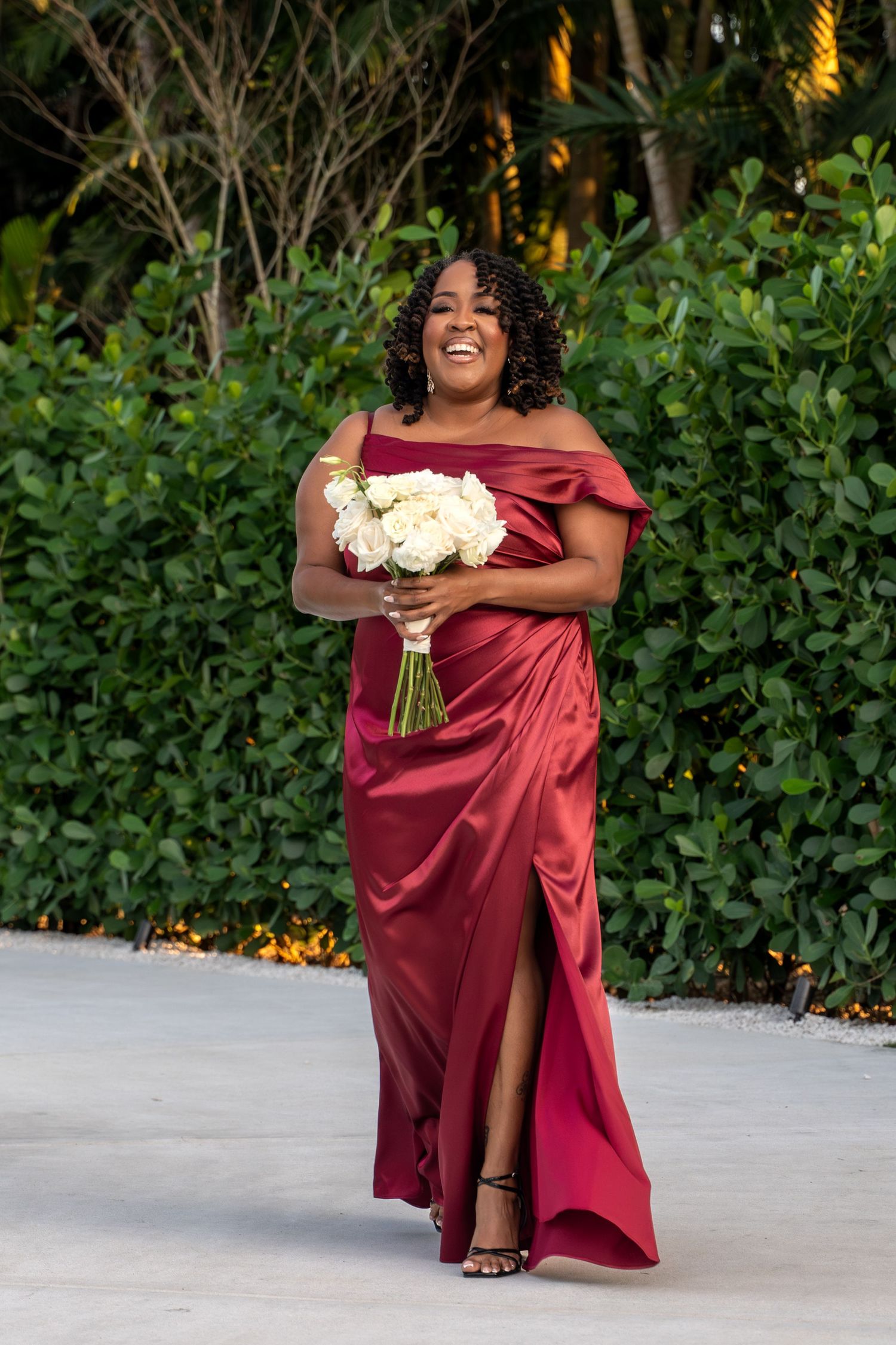 Bridesmaid in a burgundy satin dress holds a white floral bouquet while posing outdoors.