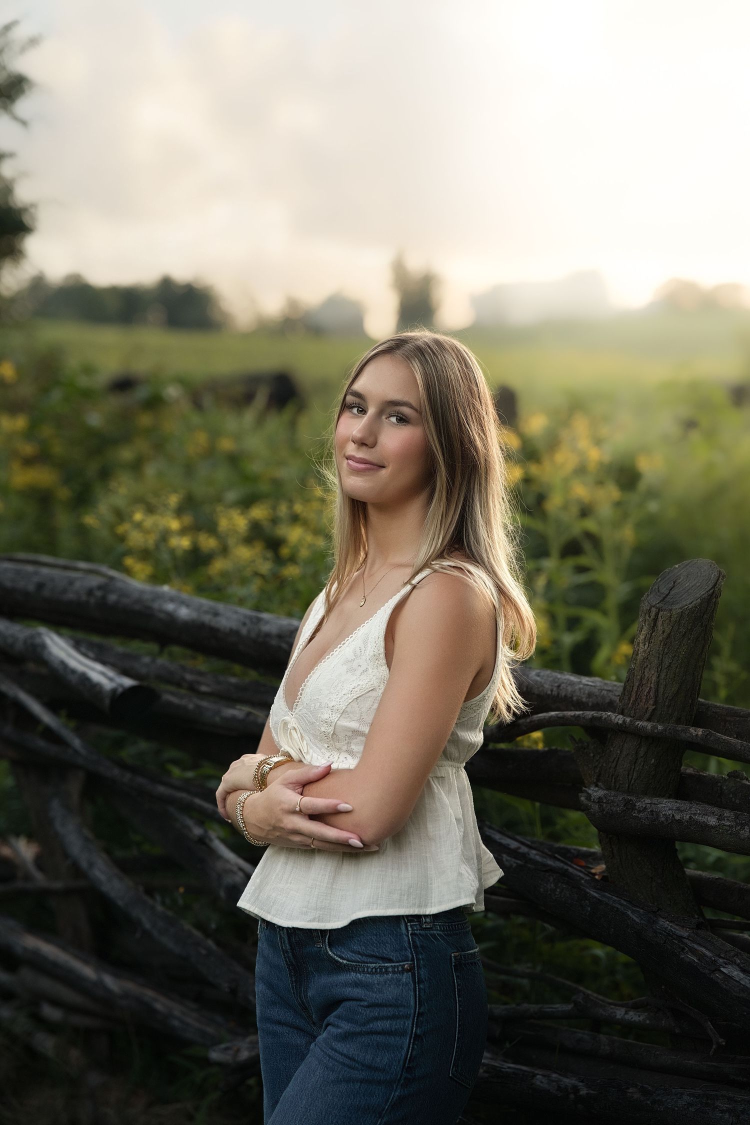 Young blonde woman in white top and jeans poses by rustic wooden fence in golden hour sunlight with green field backdrop.