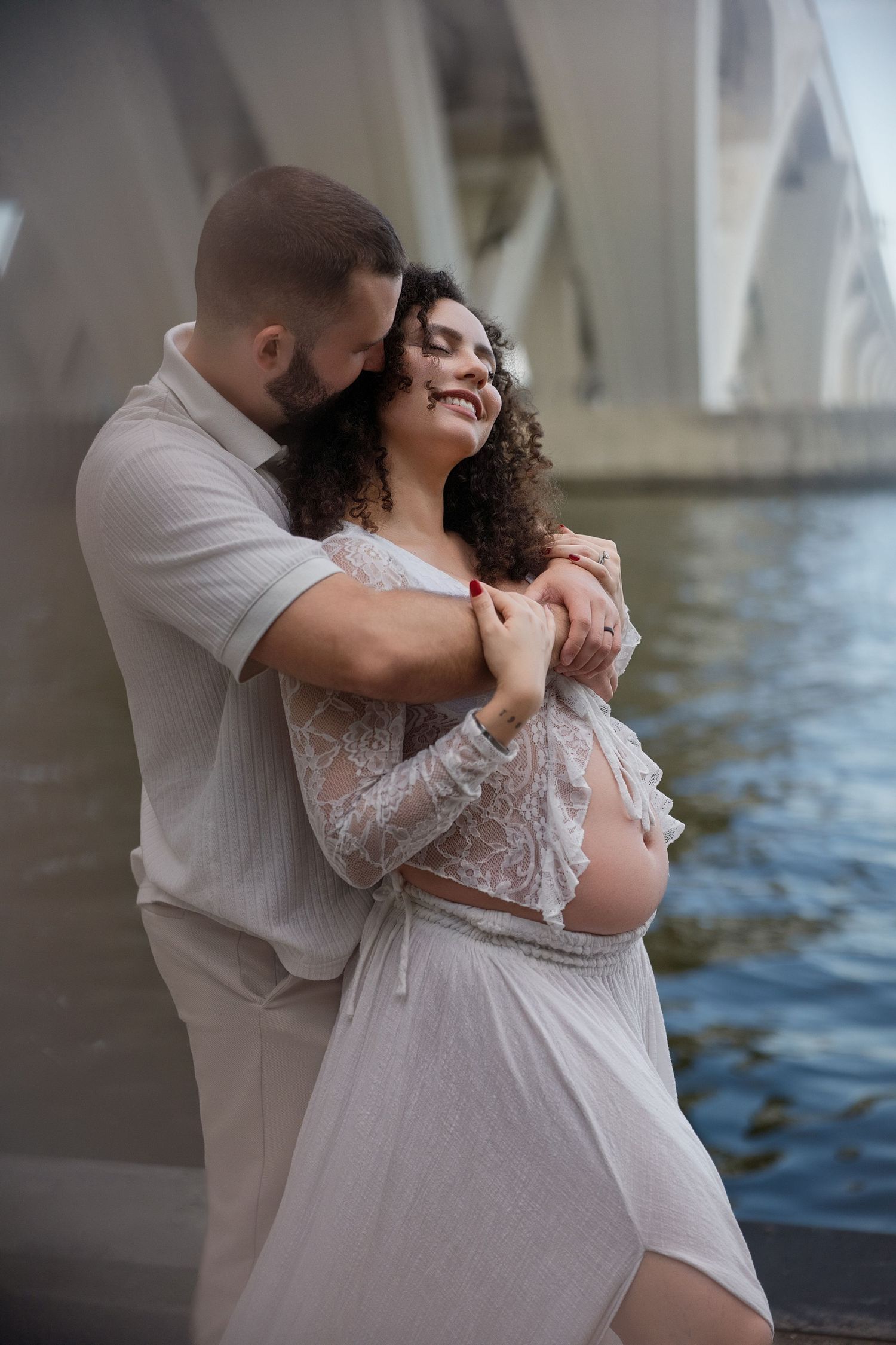 Intimate maternity couple poses under bridge architecture with water views.