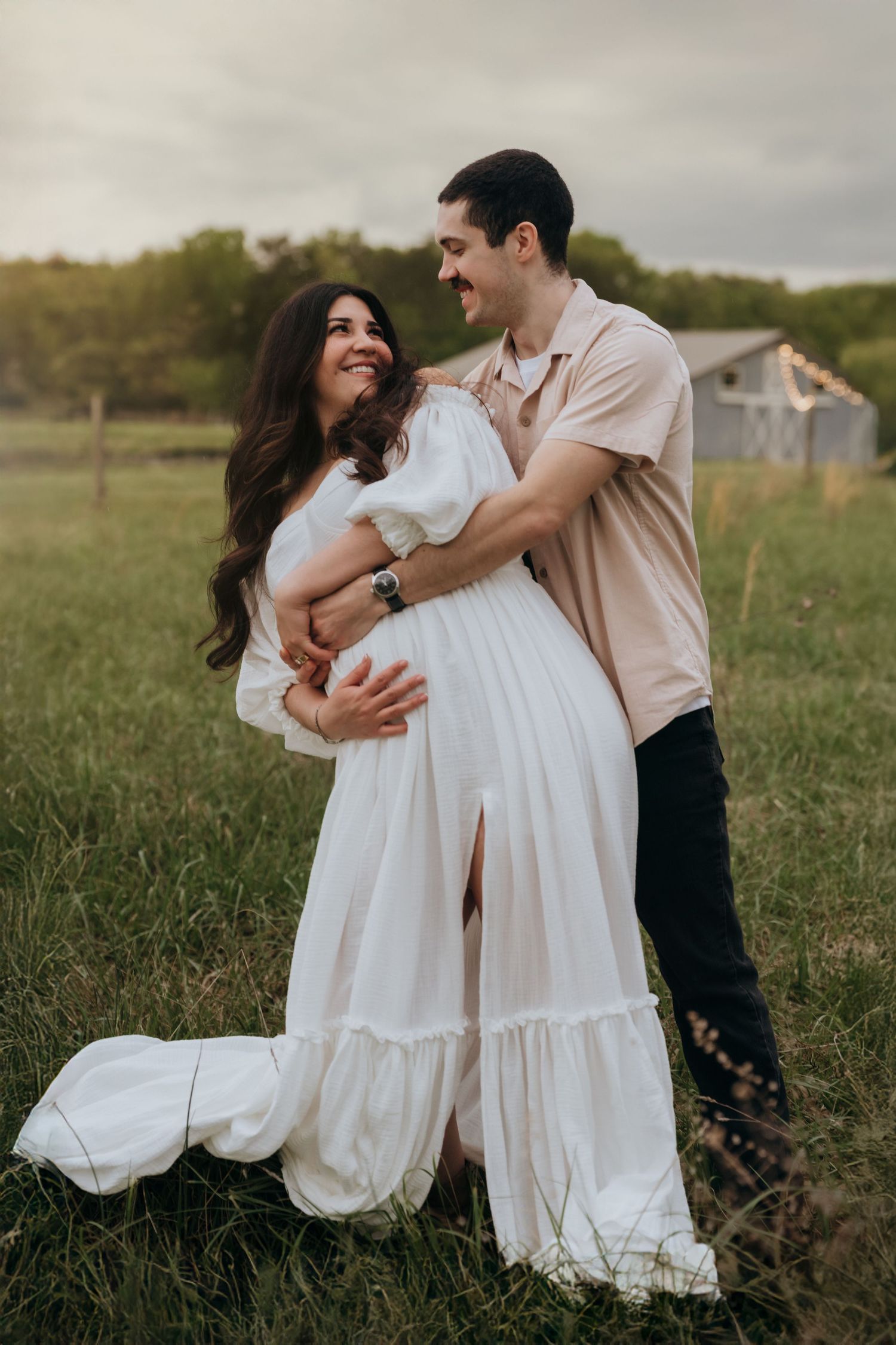 A couple in white dresses embrace lovingly in a rustic field during a romantic outdoor photoshoot.