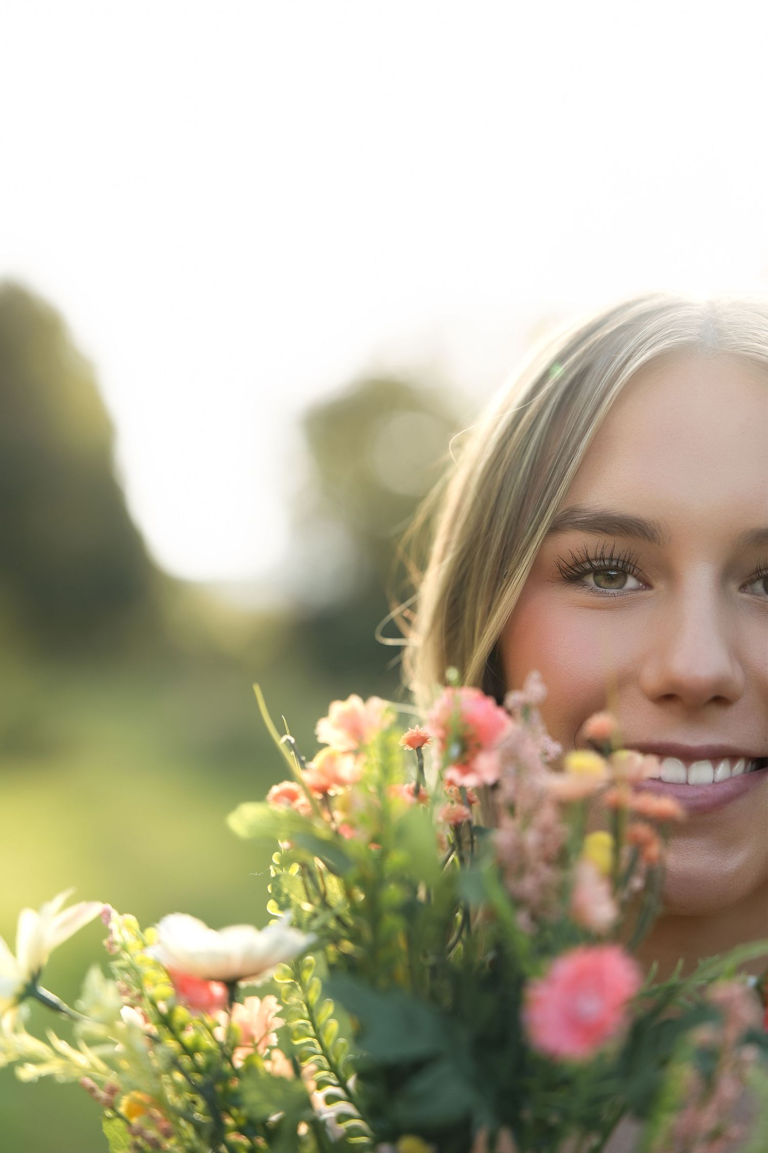 Smiling blonde woman holding a bouquet of pink and white wildflowers outdoors in soft natural sunlight.